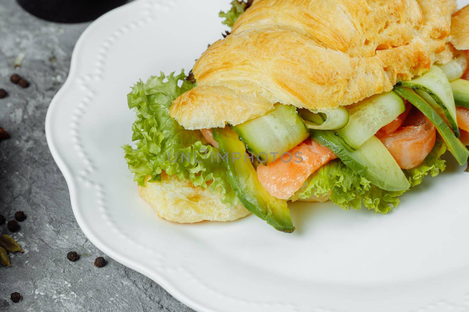 Croissant sandwich with red fish, avocado, fresh vegetables and arugula on black shale board over black stone background. Healthy food concept.