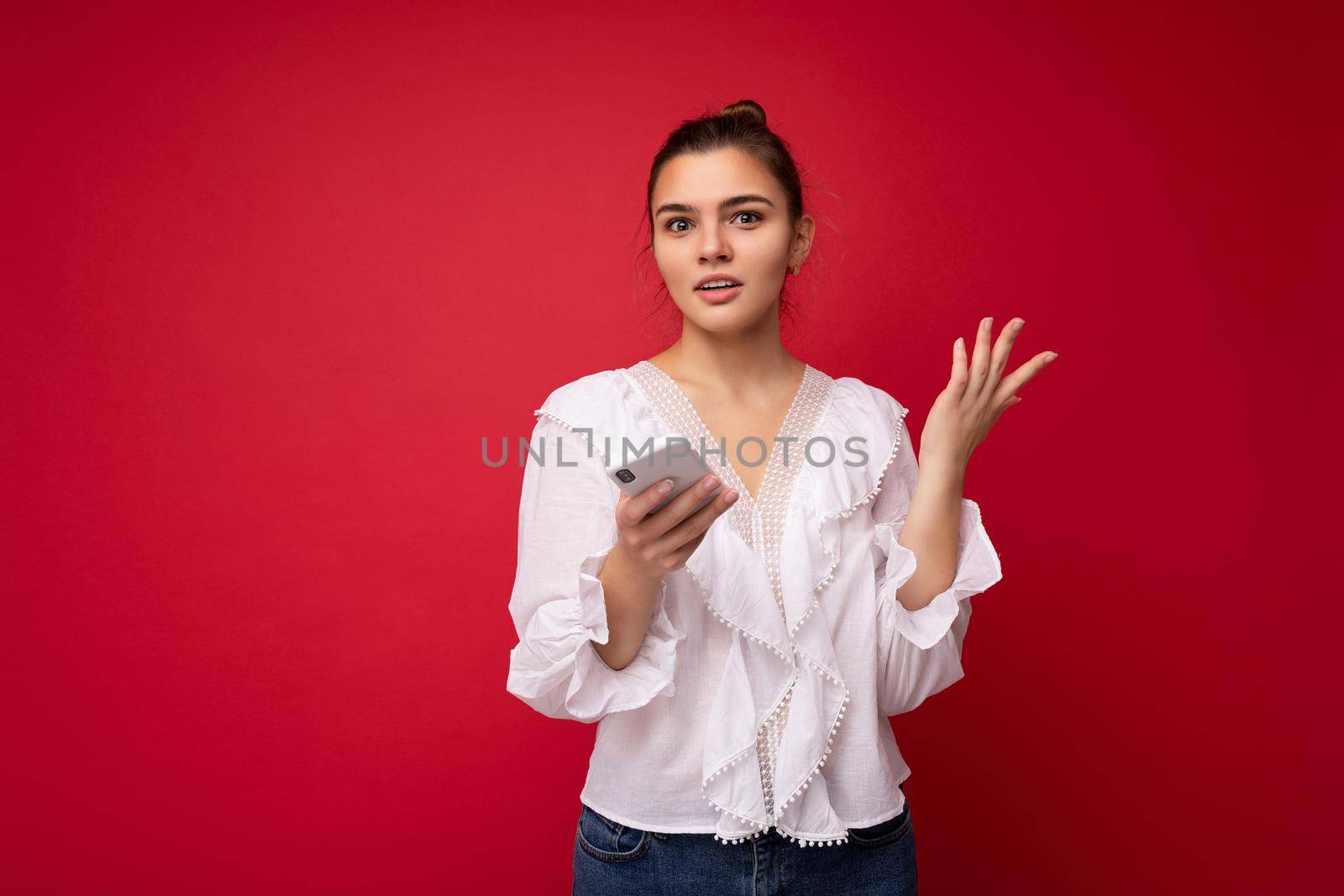 Attractive young upset brunet woman wearing white blouse standing isolated over red background reading news on the internet via phone looking at camera and don't understanding.