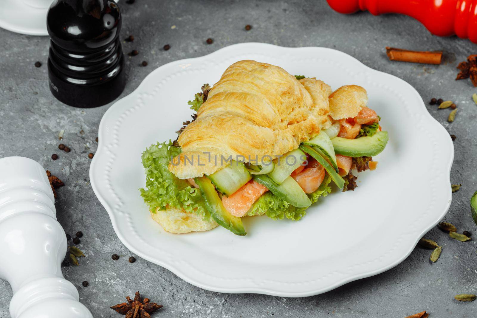 Croissant sandwich with red fish, avocado, fresh vegetables and arugula on black shale board over black stone background. Healthy food concept.