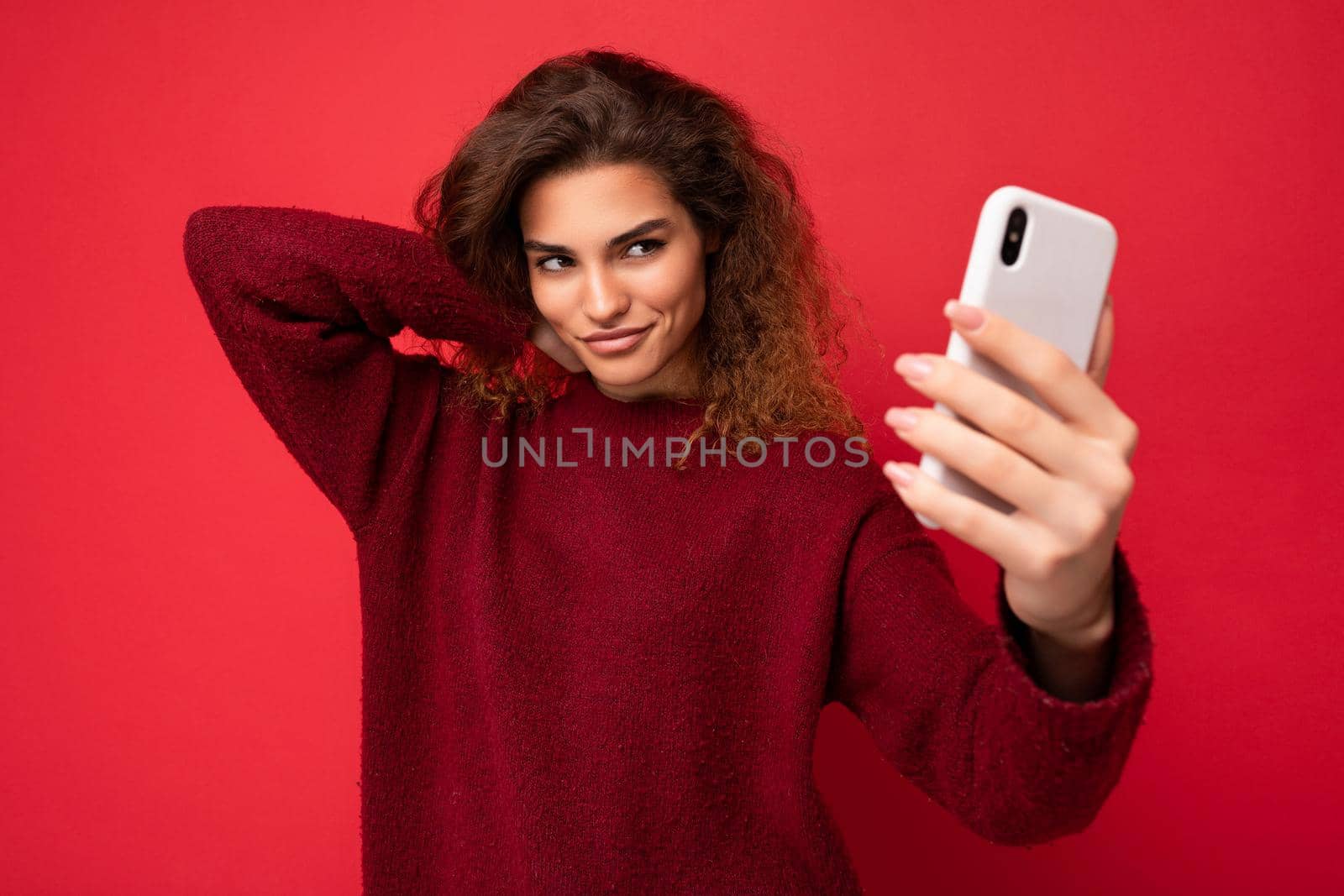 Beautiful young woman with curly hair wearing dark red sweater isolated on red background wall holding and using smart phone looking at telephone screen and taking selfie.