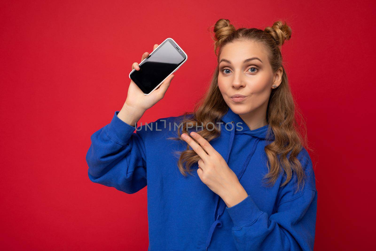 Attractive smiling young blonde woman wearing stylish blue hoodie isolated on red background with copy space holding smartphone showing phone in hand with empty screen display looking at camera showing.