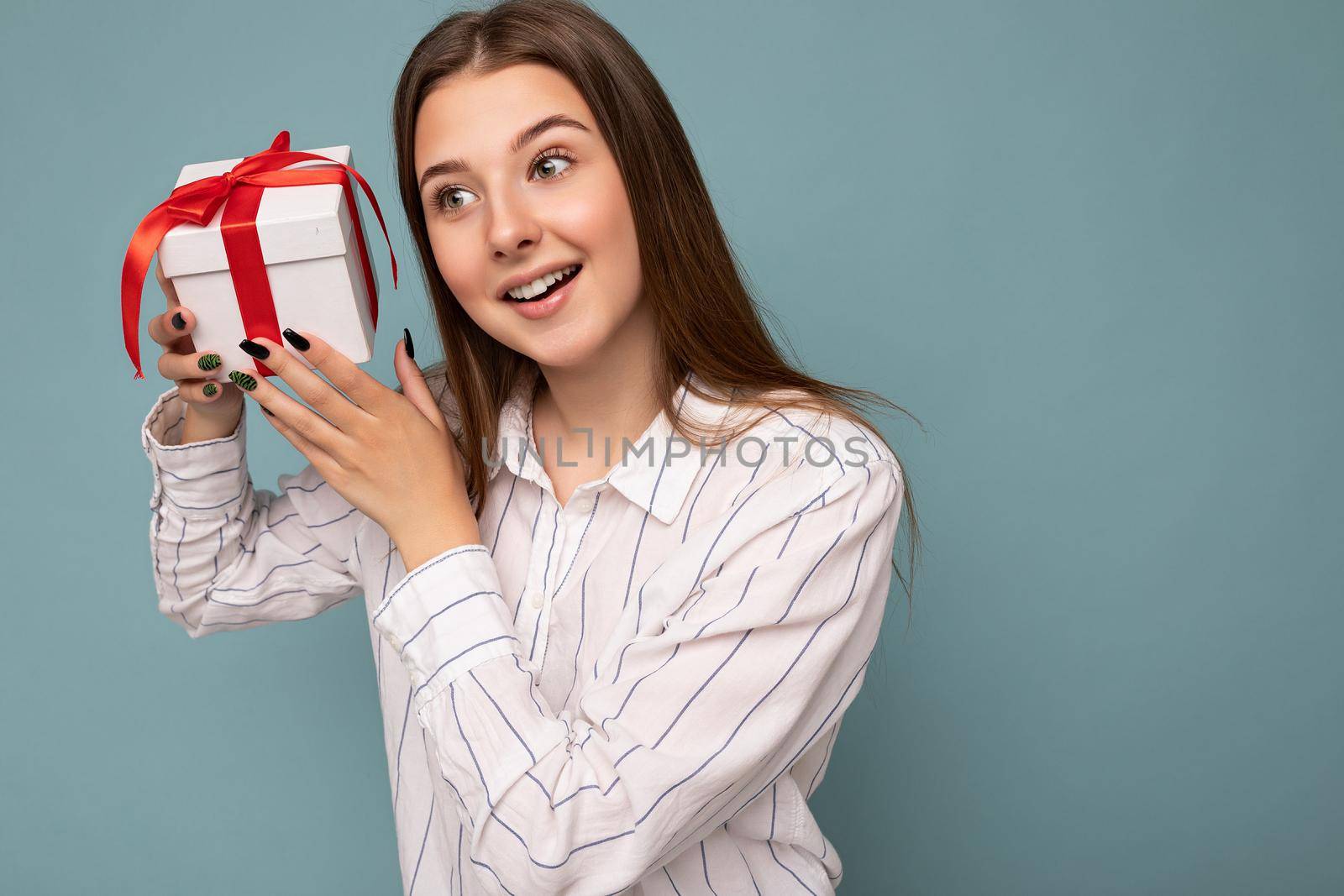 Photo of charming happy positive surprised brunette young woman isolated over blue background wall wearing white shirt holding white gift box with red ribbon and looking to the side. Copy space
