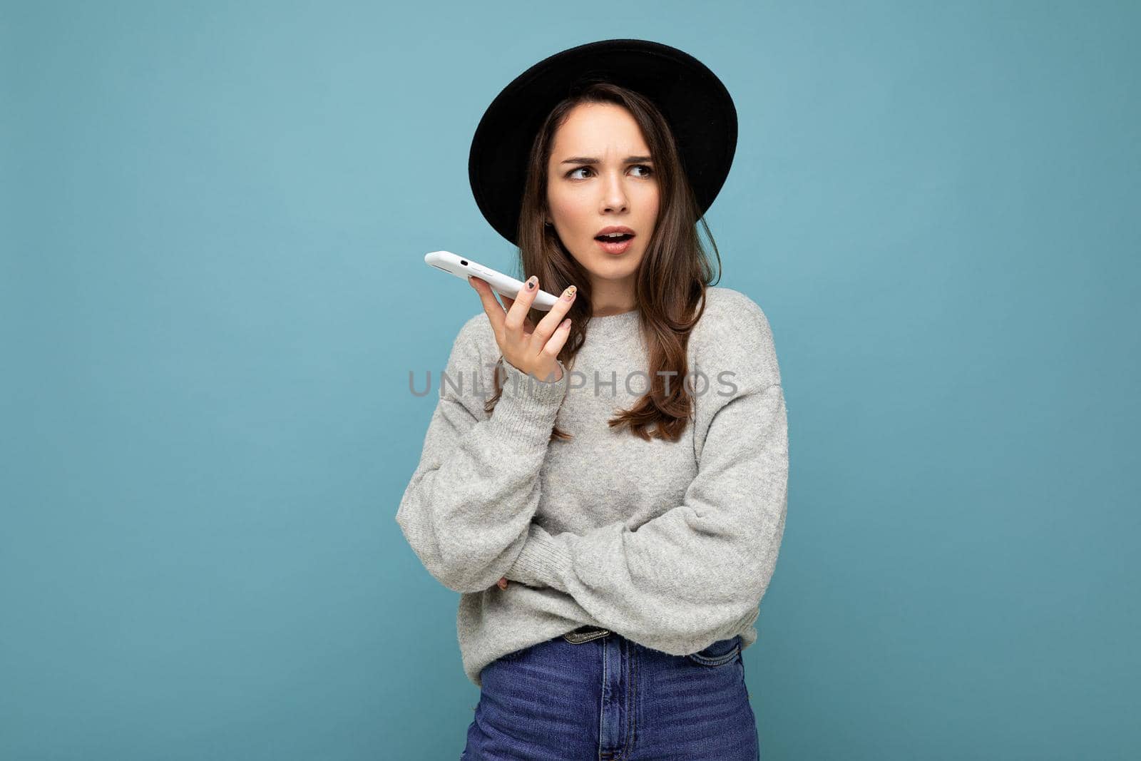 Beautiful young brunette woman thinking wearing black hat and grey sweater holding smartphone looking to the side texting isolated on background.