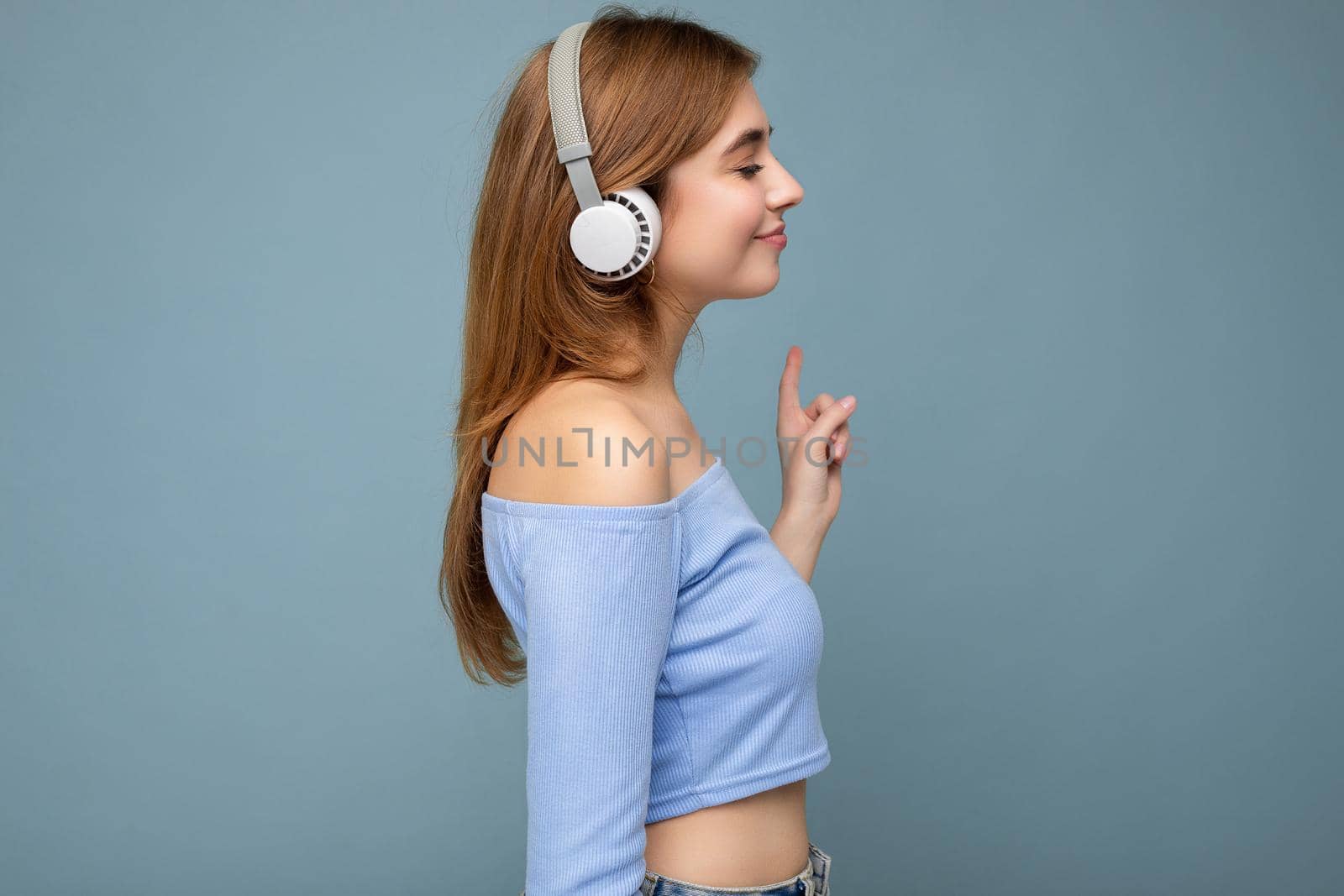 Side-profile photo of beautiful positive smiling young blonde female person wearing blue crop top isolated over blue background wall wearing white wireless bluetooth headphones listening to cool music and looking to the side.
