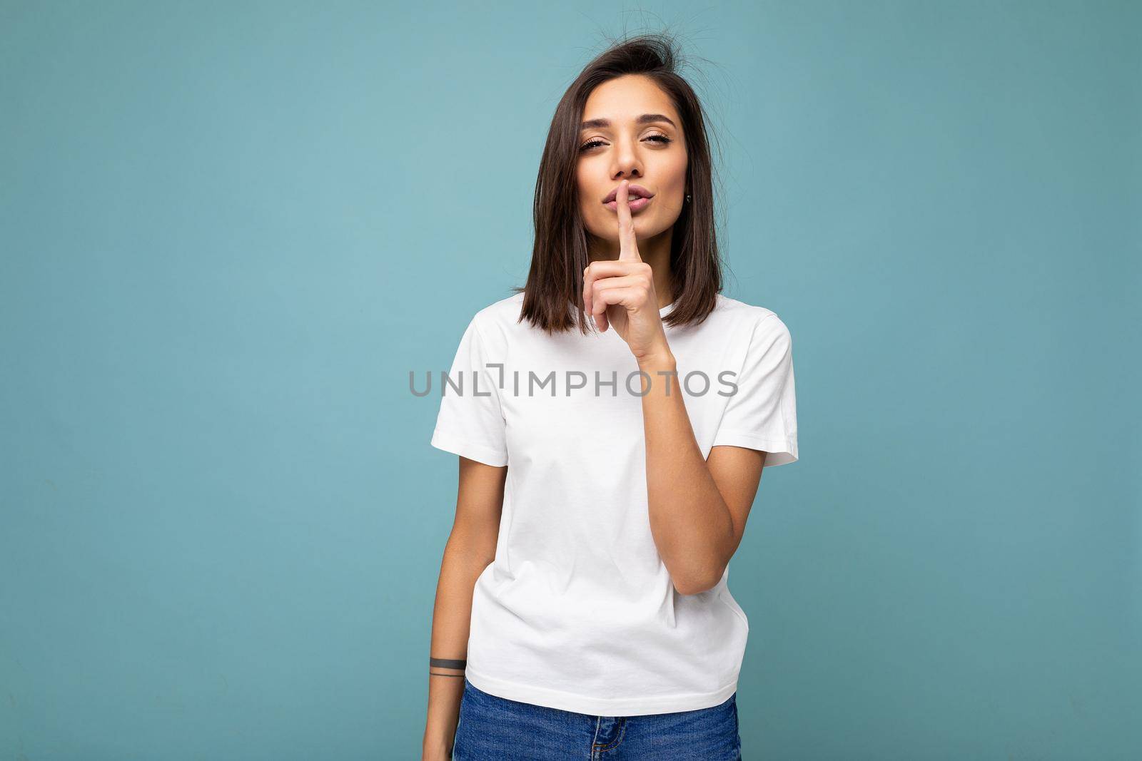 Portrait of young attractive brunette woman with sincere emotions wearing casual white t-shirt for mockup isolated on blue background with copy space and showing shh gesture. Keep secret.