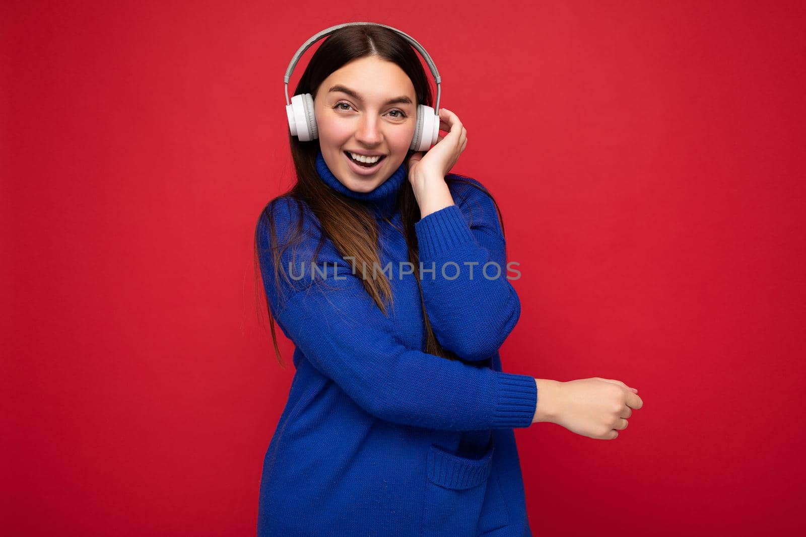 Attractive positive young brunette woman wearing blue sweater isolated over red background wall wearing white bluetooth earphones listening to cool music and having fun looking at camera.