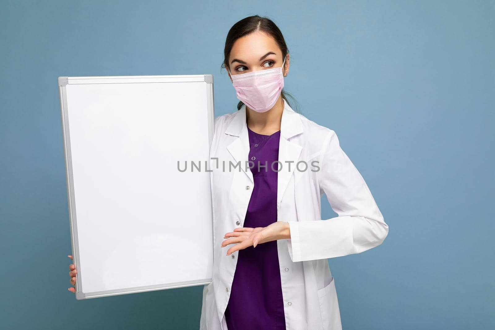 Beautiful brunette female nurse in protective face mask and white medical coat holding an empty magnetic board isolated on blue background.