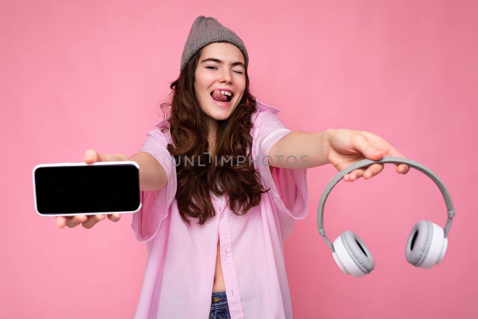 Photo of beautiful positive smiling young woman wearing stylish casual outfit isolated on colorful background wall holding white bluetooth wireless earphones and showing mobile phone with empty screen for mockup looking at camera.