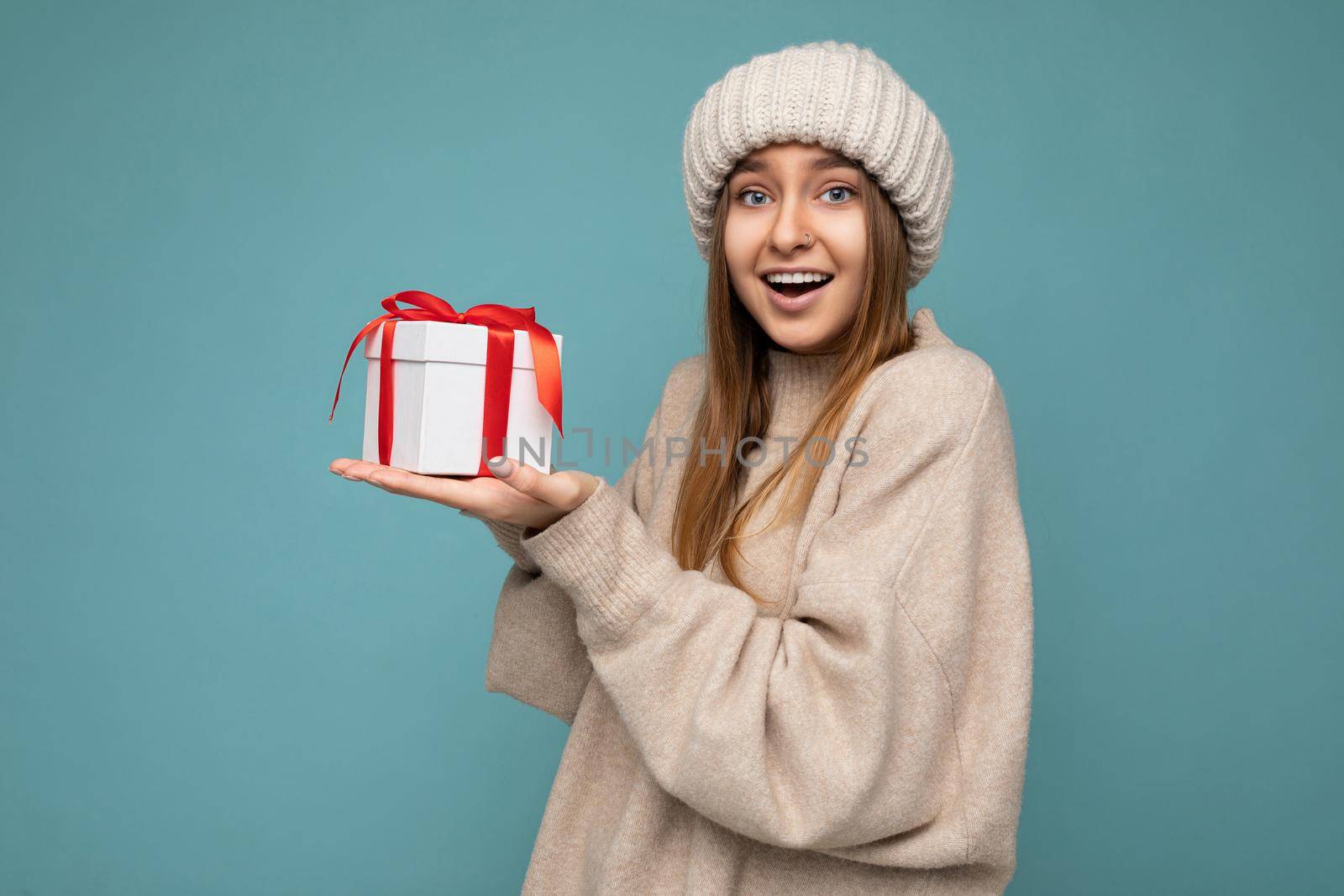 Photo shot of pretty positive surprised young dark blond woman isolated over colourful background wall wearing trendy winter outfit holding gift box and looking at camera.