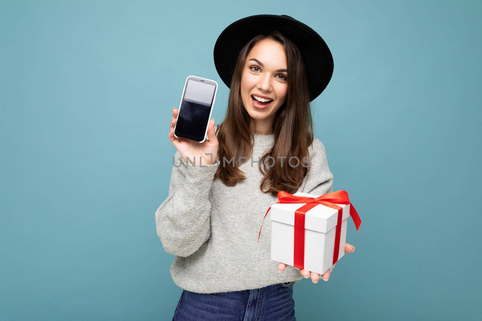 Shot of pretty smiling positive young brunette woman isolated over blue background wall wearing stylish black hat and grey sweater holding gift box showing smartphone screen display for mockup and looking at camera.