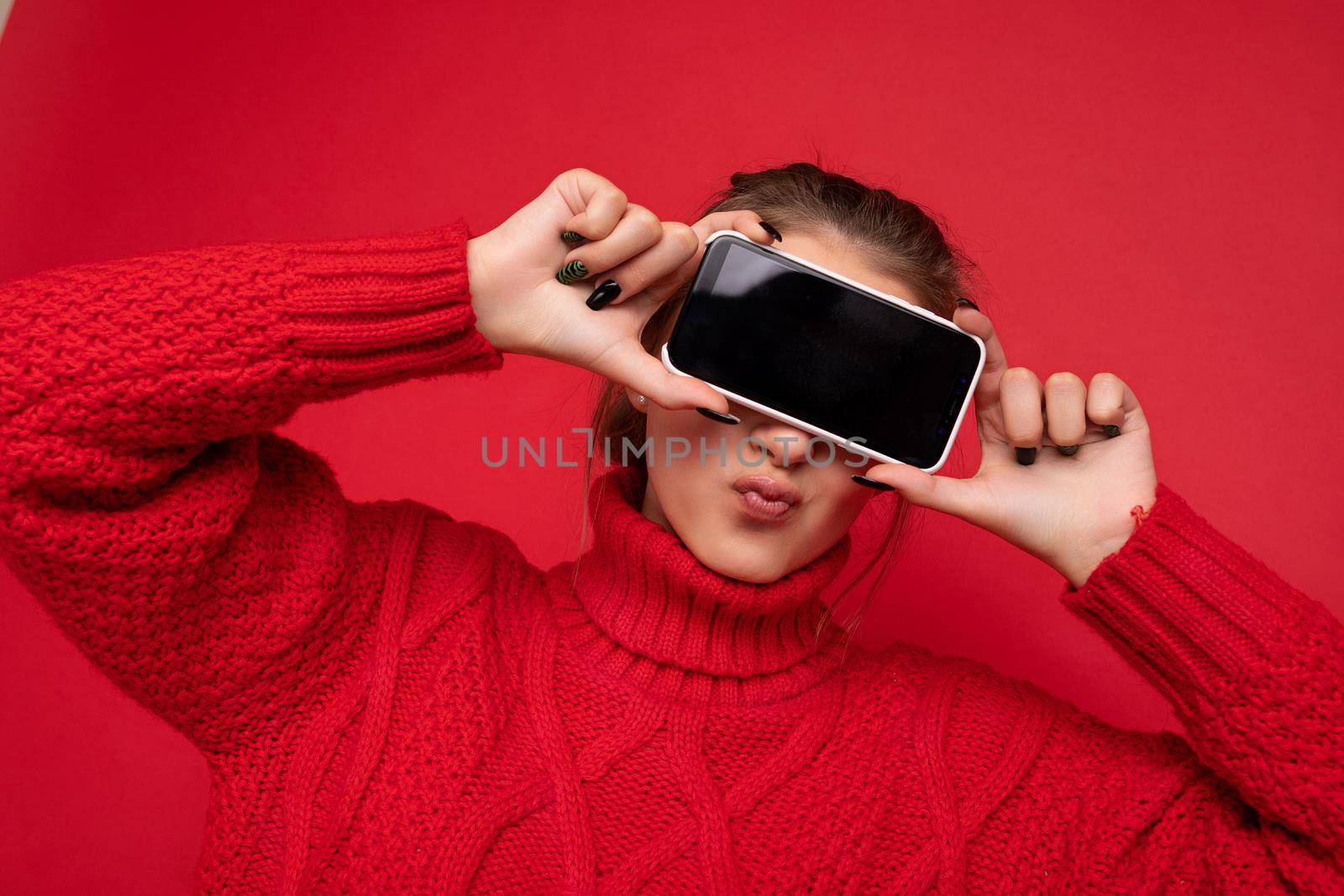 Photo of beautiful cute smiling young woman wearing warm red sweater isolated over red background wall holding smartphone and showing phone with empty display for mockup.