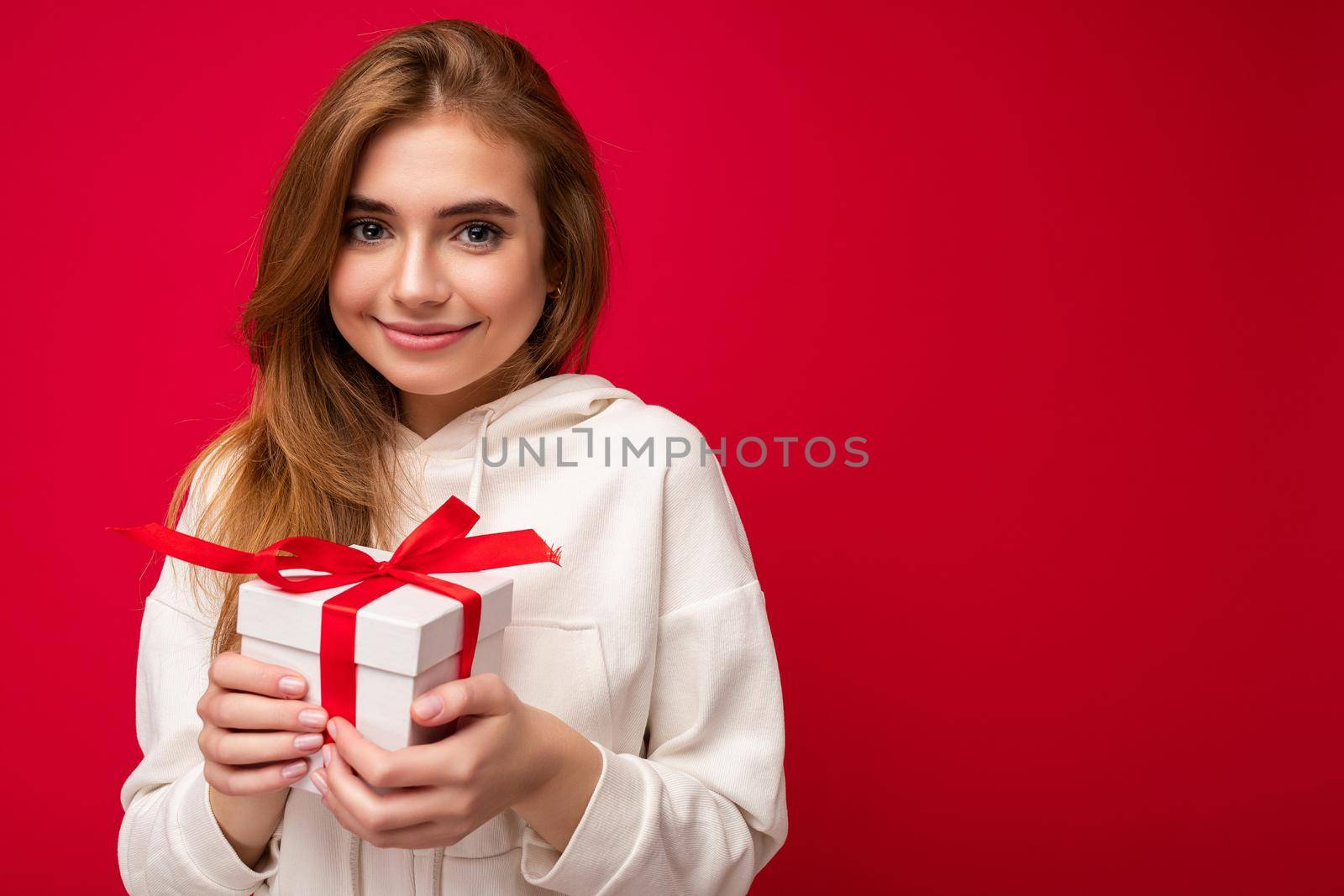 Beautiful happy young blonde woman isolated over colourful background wall wearing stylish casual clothes holding gift box and looking at camera. Copy space