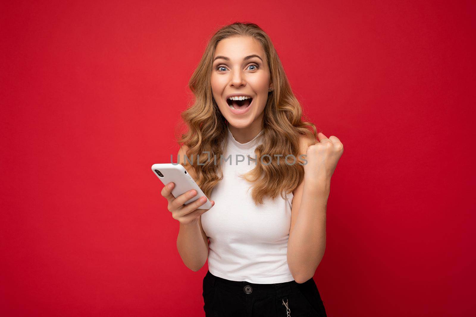 Photo of attractive crazy amazed surprised young woman wearing casual stylish clothes standing isolated over background with copy space holding and using mobile phone looking at camera.