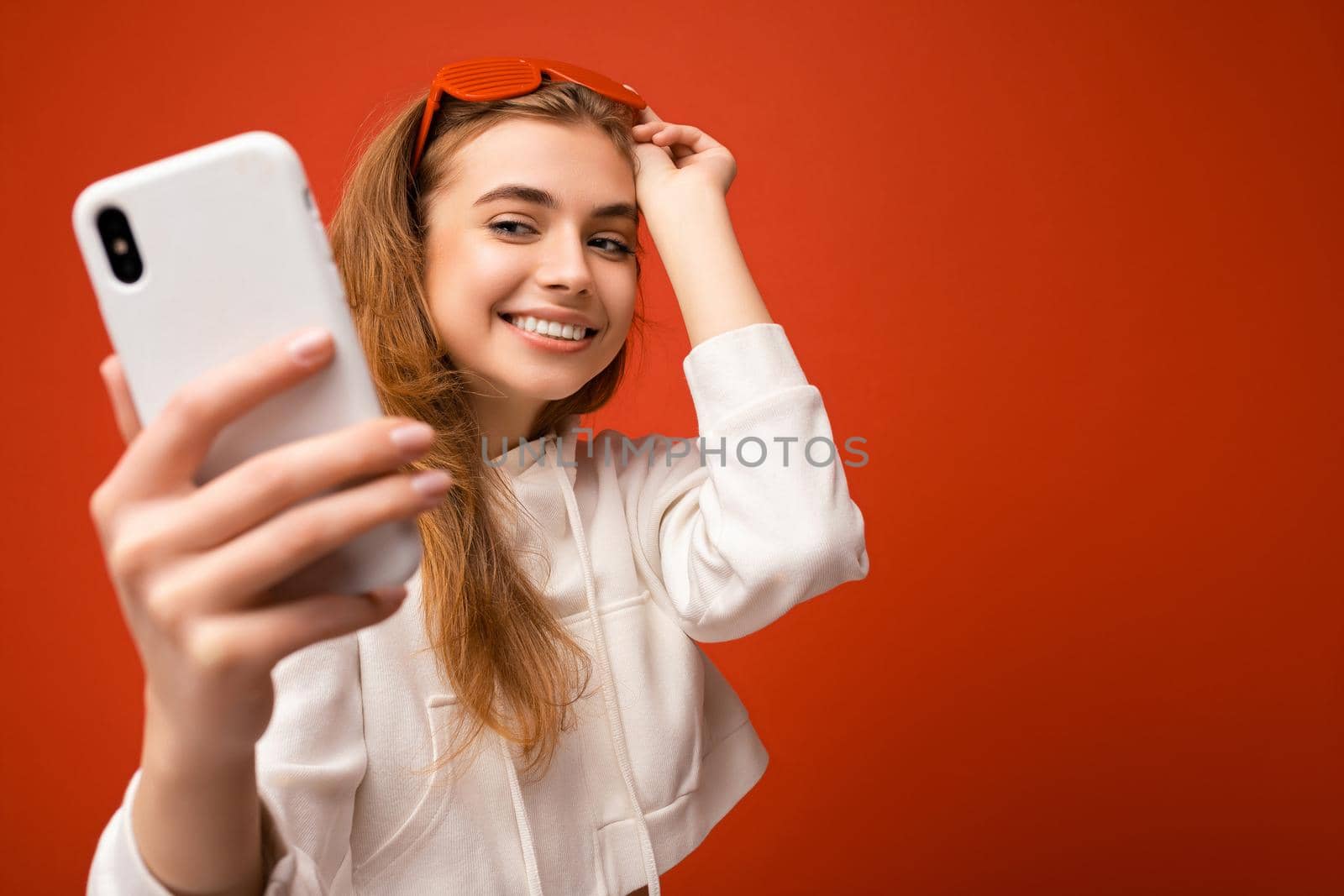 Closeup portrait of sexy attractive positive smiling young blonde woman wearing stylish white hoodie and funny colorful glasses standing isolated over colourful background holding and using mobile phone taking selfie photo looking at gadjet. Copy space