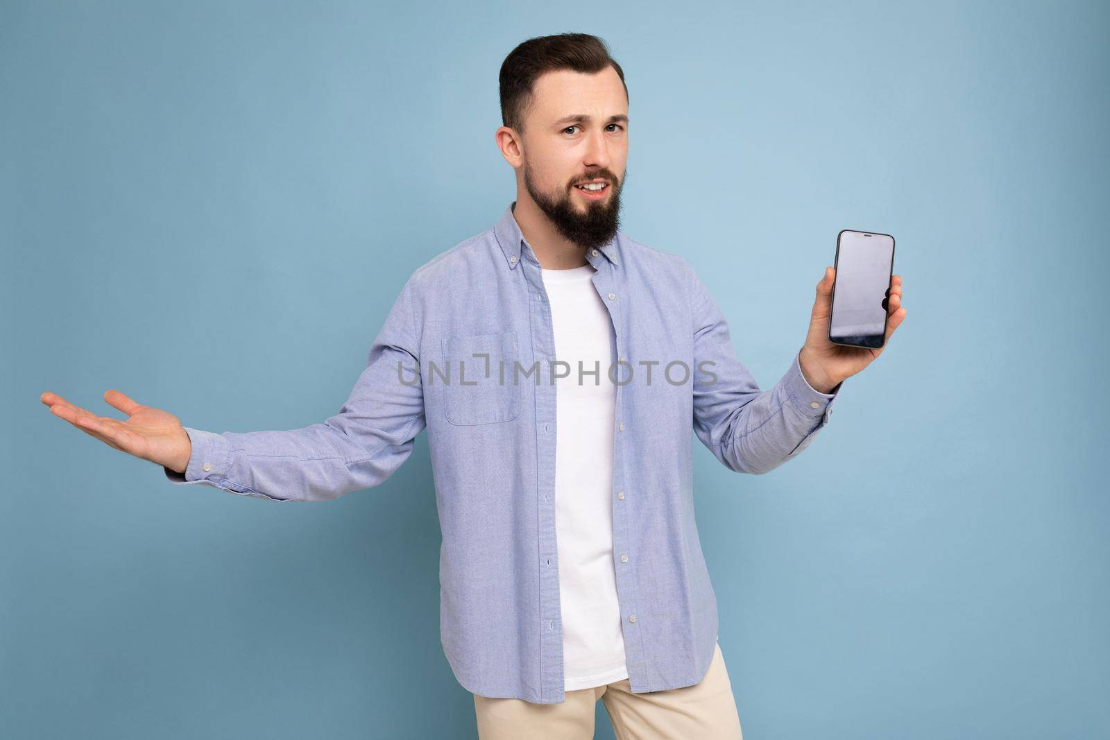 Photo of thoughtful handsome good looking young brunette unshaven man with beard wearing casual white t-shirt and blue shirt poising isolated on blue background with empty space holding in hand mobile with empty screen looking at camera and don't understanding.