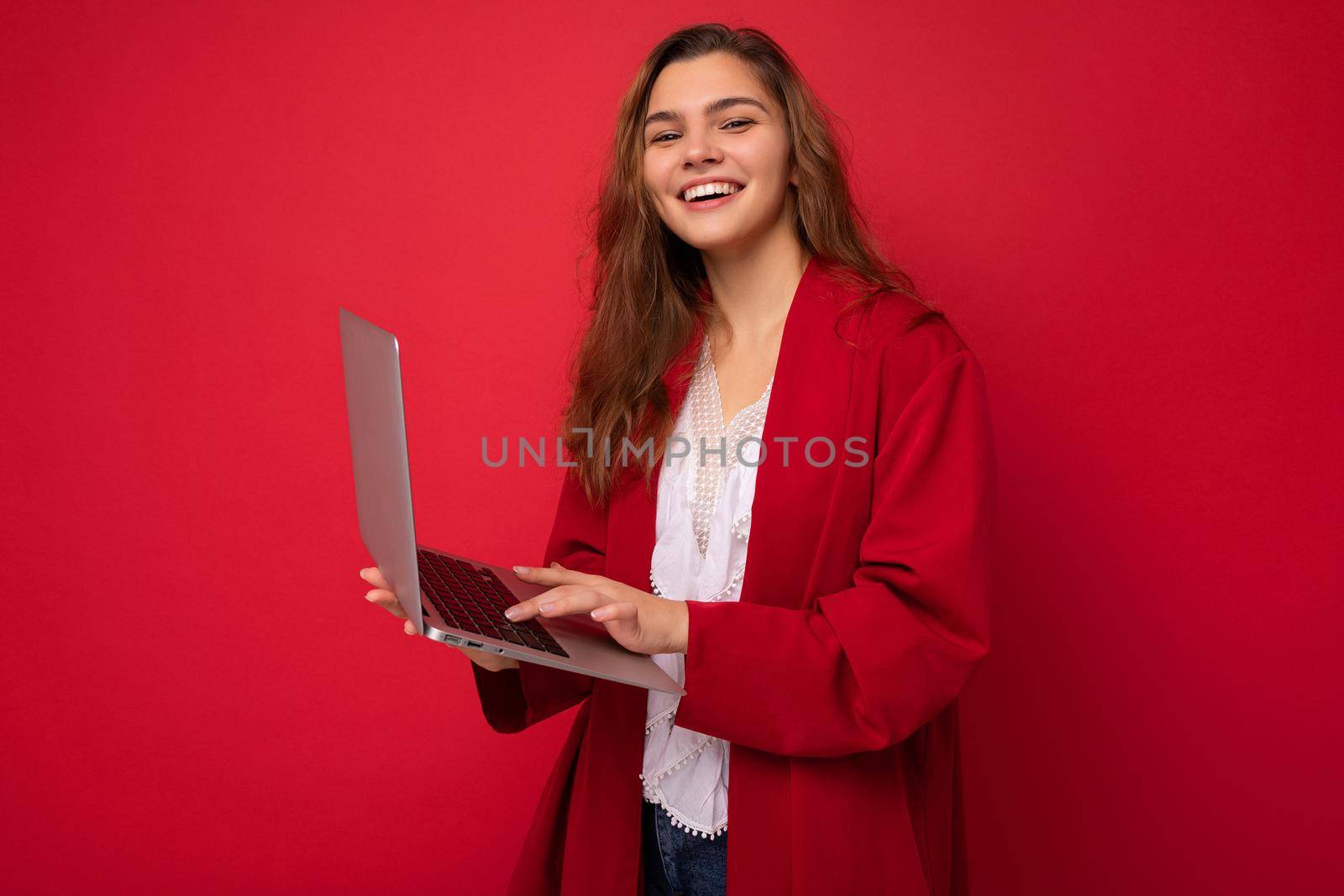 Shot fascinating smiling young brunet woman holding computer typing on keyboard looking at camera wearing red cardigan and white t-shirt isolated on red wall background.