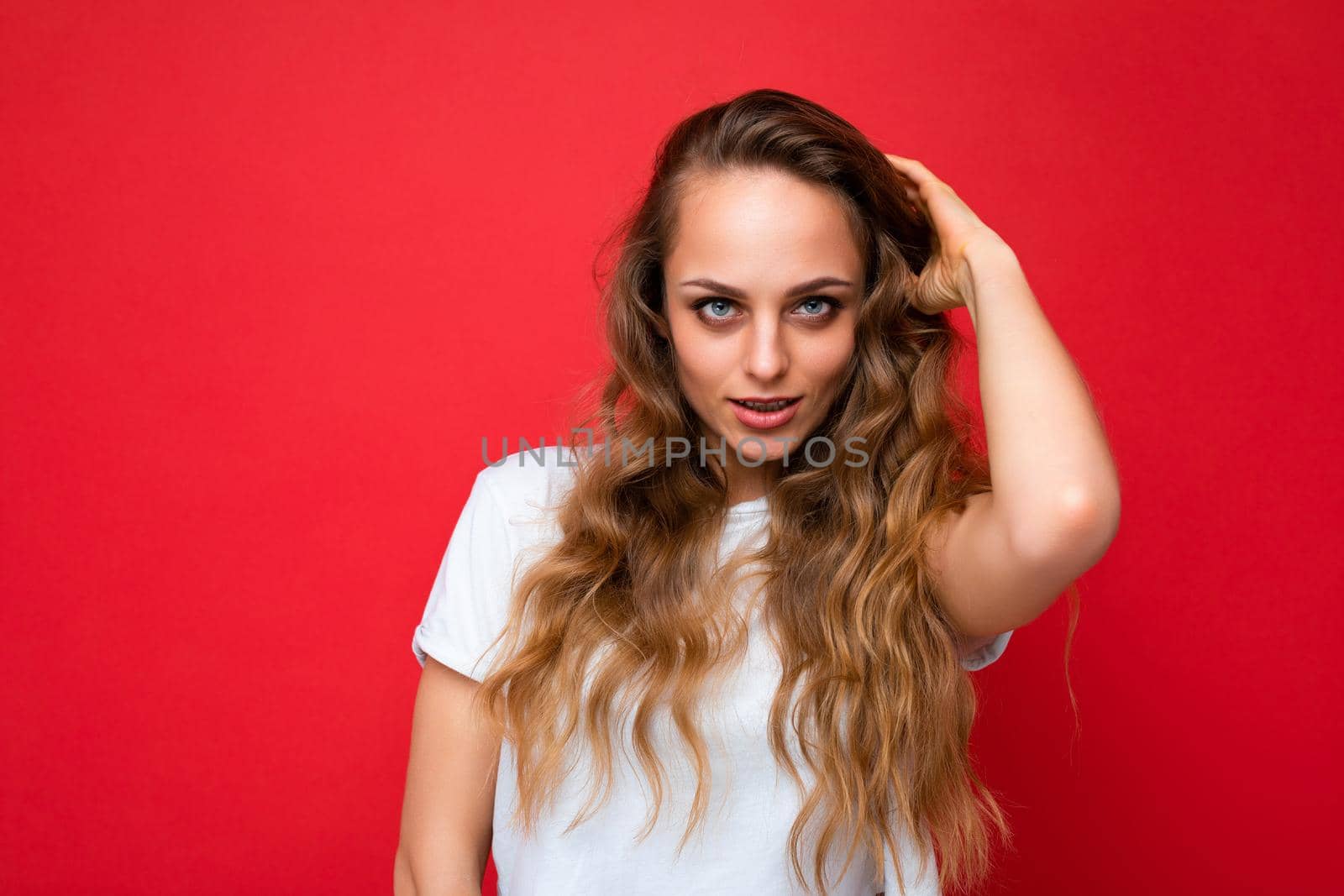 Young beautiful curly blonde woman with sexy expression, cheerful and happy face wearing casual white t-shirt isolated over red background with copy space.