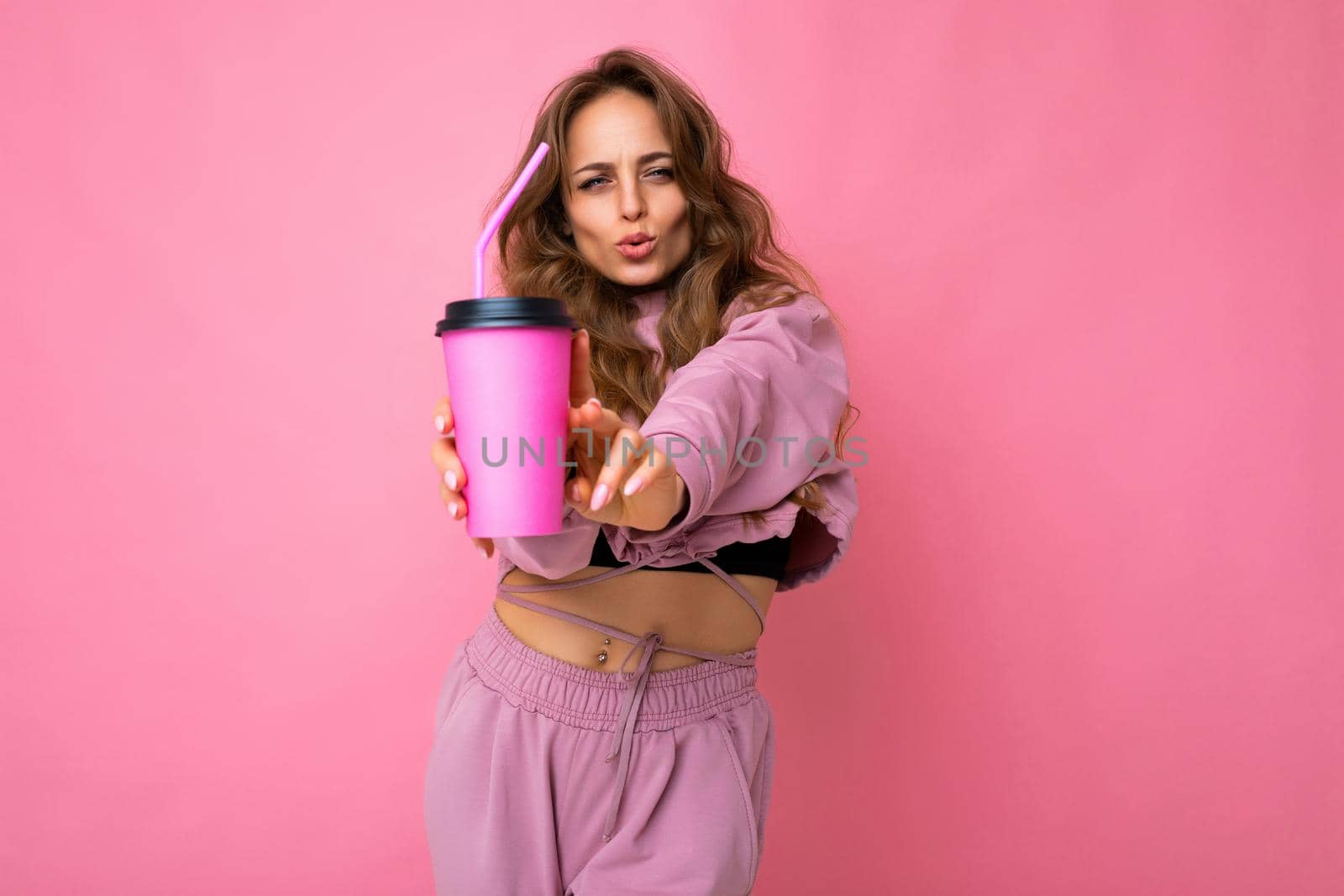 Photo shot of beautiful adult happy funny blonde female person wearing pink sport clothes isolated over pink background holding and suggesting paper coffee cup for mockup drinking and looking at camera.