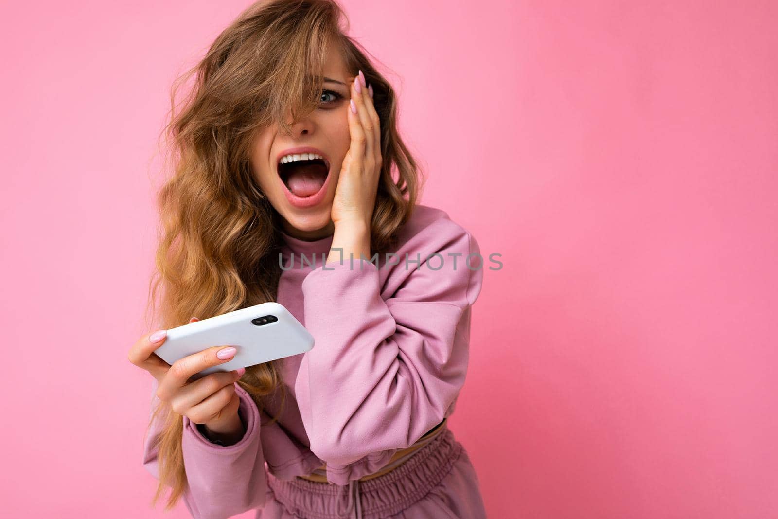 Photo of attractive crazy amazed surprised young woman wearing casual stylish clothes standing isolated over background with copy space holding and using mobile phone looking at camera and shouting.