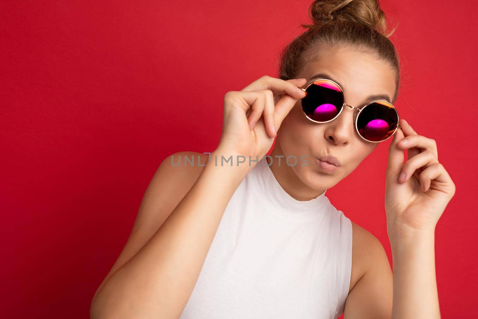 Photo shot of beautiful young blonde woman wearing casual clothes and stylish sunglasses isolated over colorful background looking at camera and giving kiss.