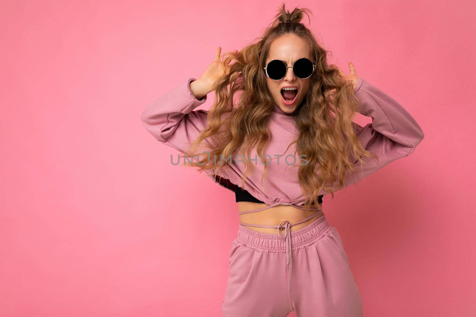 Shot of beautiful happy positive joyful young dark blonde curly woman isolated over pink background wall wearing casual pink sport clothes and stylish sunglasses looking at camera and dancing. copy space