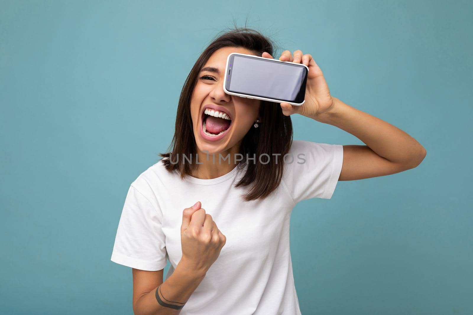 Attractive smiling young brunet woman good looking wearing white t-shirt standing isolated on blue background with copy space holding smartphone showing phone in hand with empty screen screen for cutout shouting showing yes gesture.