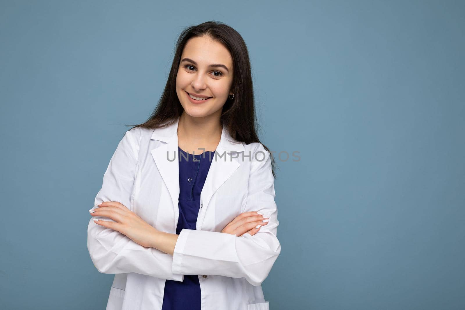 Photo portrait of young pretty beautiful positive smiling brunette woman with sincere emotions wearing white medical coat isolated over blue background with copy space and holding crossed arms.