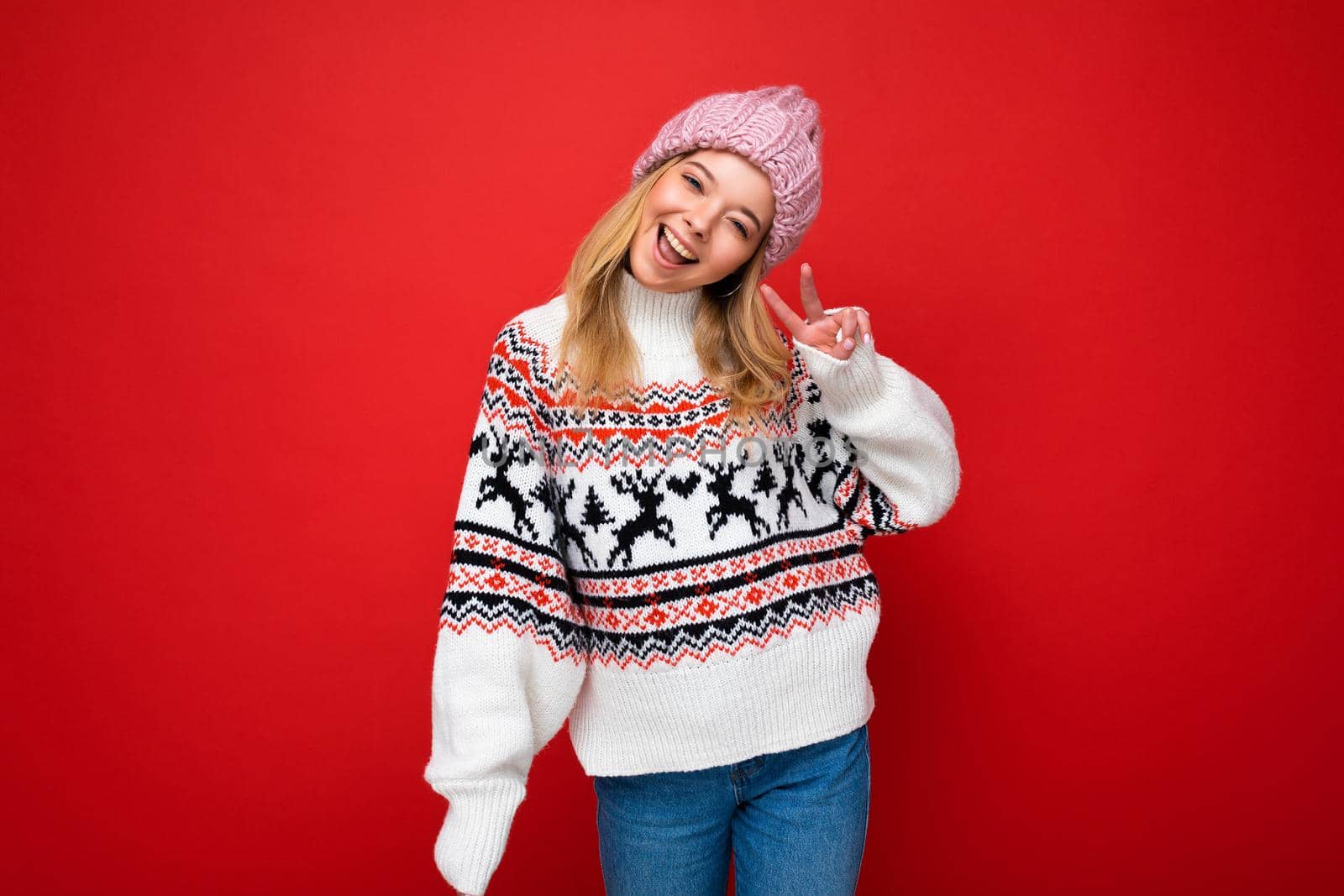 Photo of young positive happy beautiful blonde woman with sincere emotions wearing pink knitted hat and winter pullover isolated over red background with empty space and showing peace gesture by TRMK