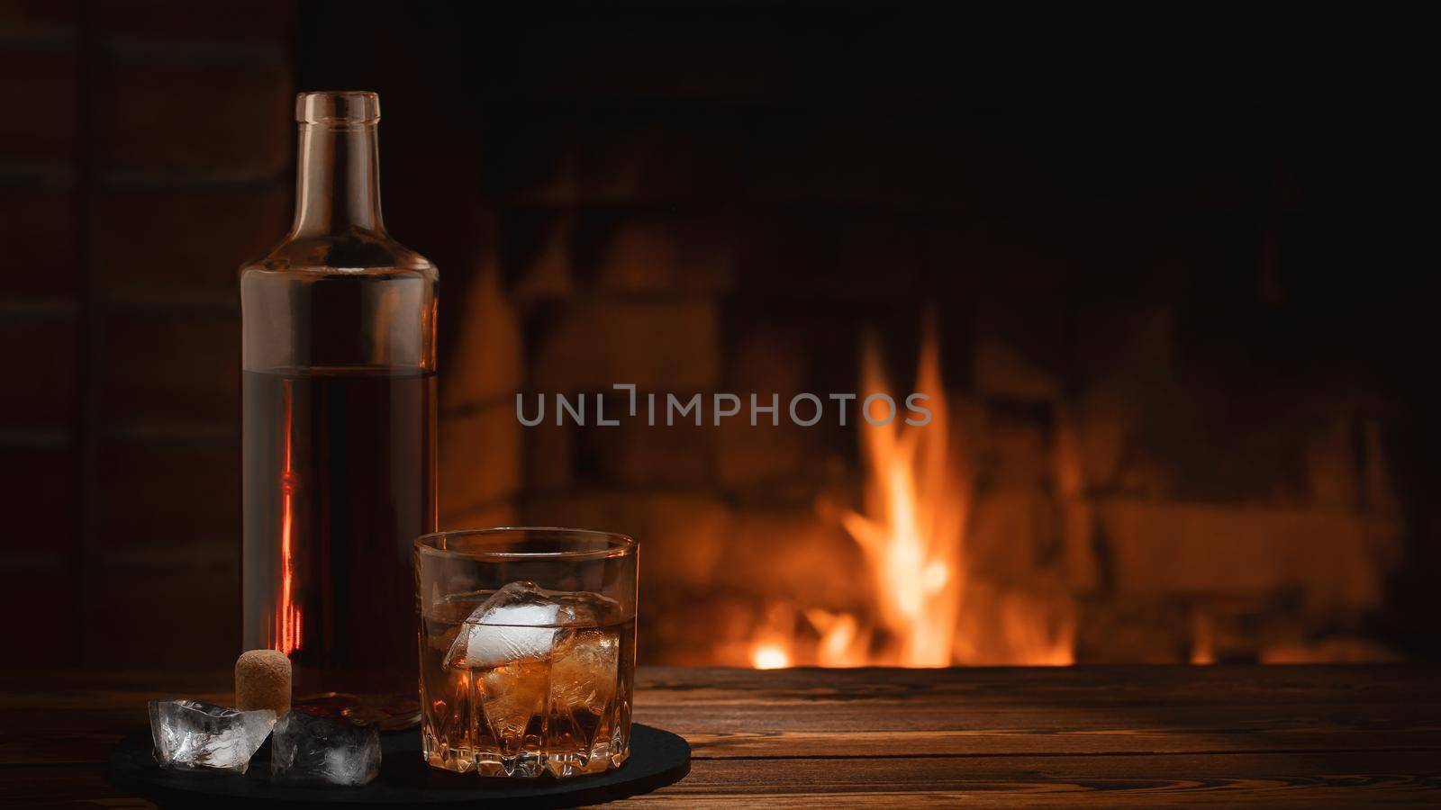 Bottle and glass of whiskey with ice on the table near the burning fireplace. Rest and relaxation concept.