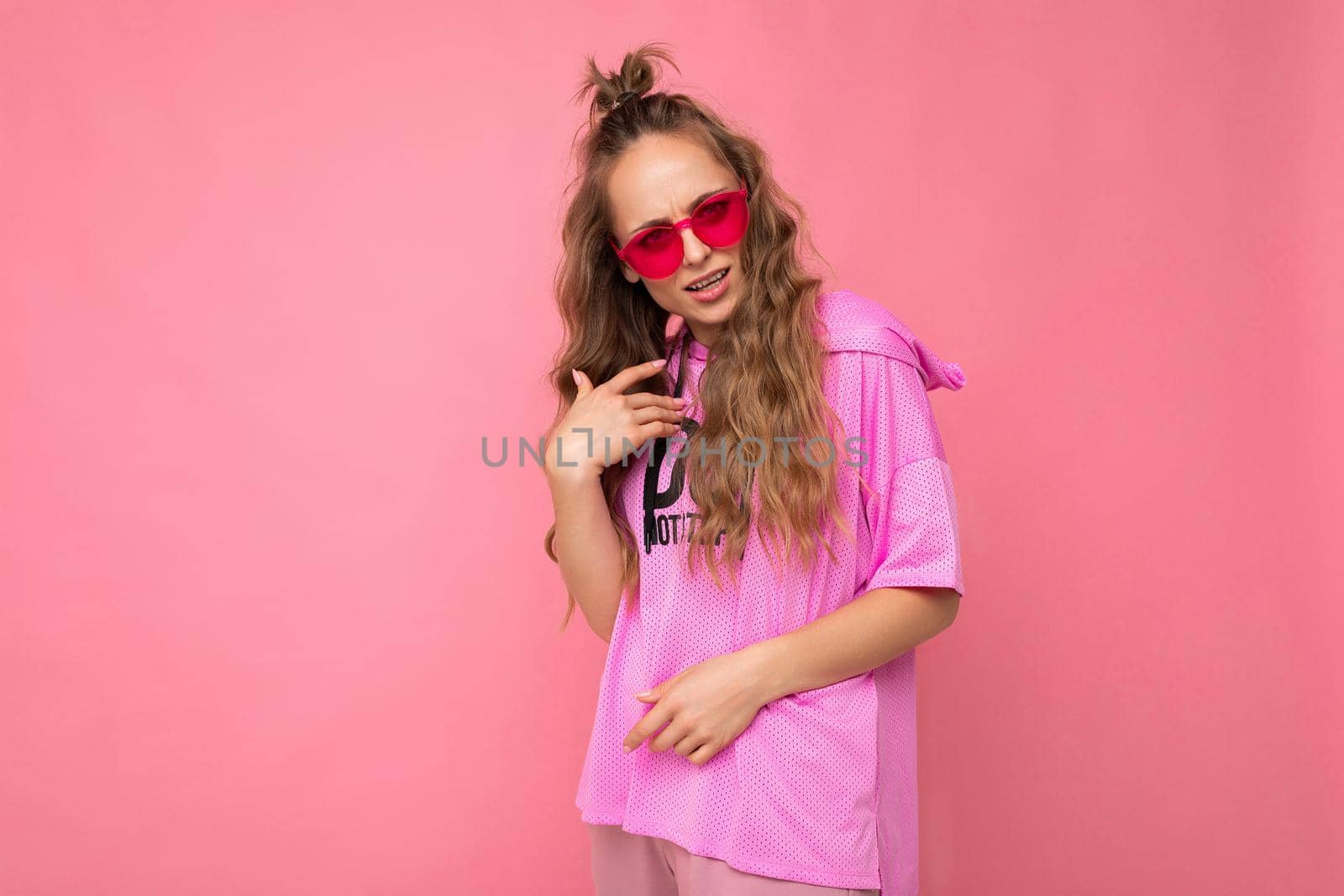 Charming sad young blonde curly woman isolated on pink background wall wearing casual pink t-shirt and stylish colourful sunglasses looking at camera and asking.