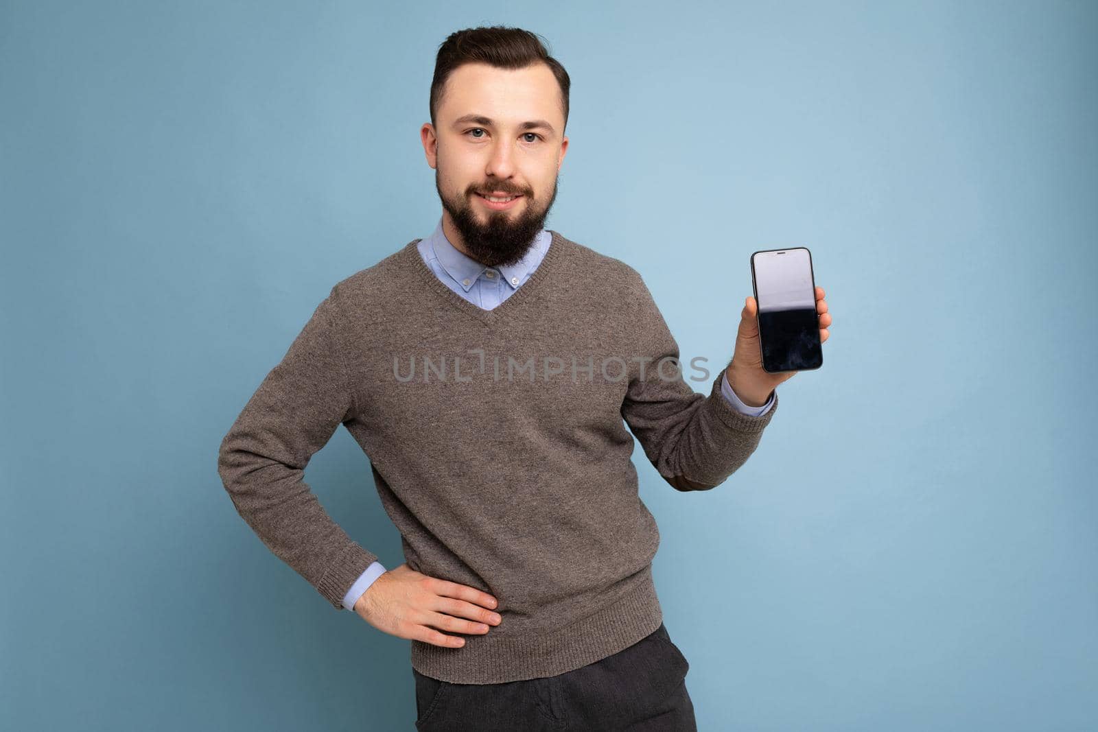 Handsome happy cool young brunette unshaven man with beardwearing stylish grey sweater and blue shirt standing isolated over pink background wall holding smartphone and showing phone with empty screen display looking at camera.