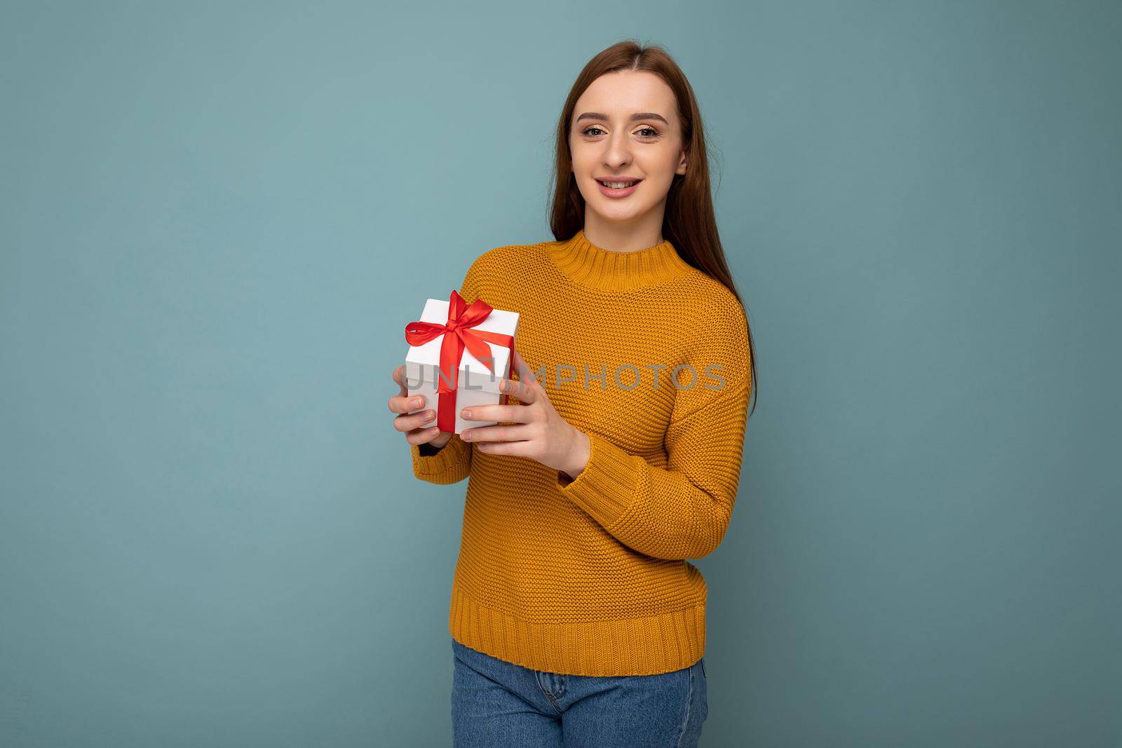 Beautiful happy young brunette woman isolated over colourful background wall wearing stylish casual clothes holding gift box and looking at camera.