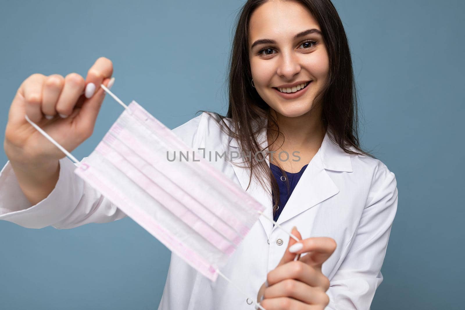 Happy smiling woman holding protective mask and wearing white medical coat isolated on blue background. Coronavirus concept.