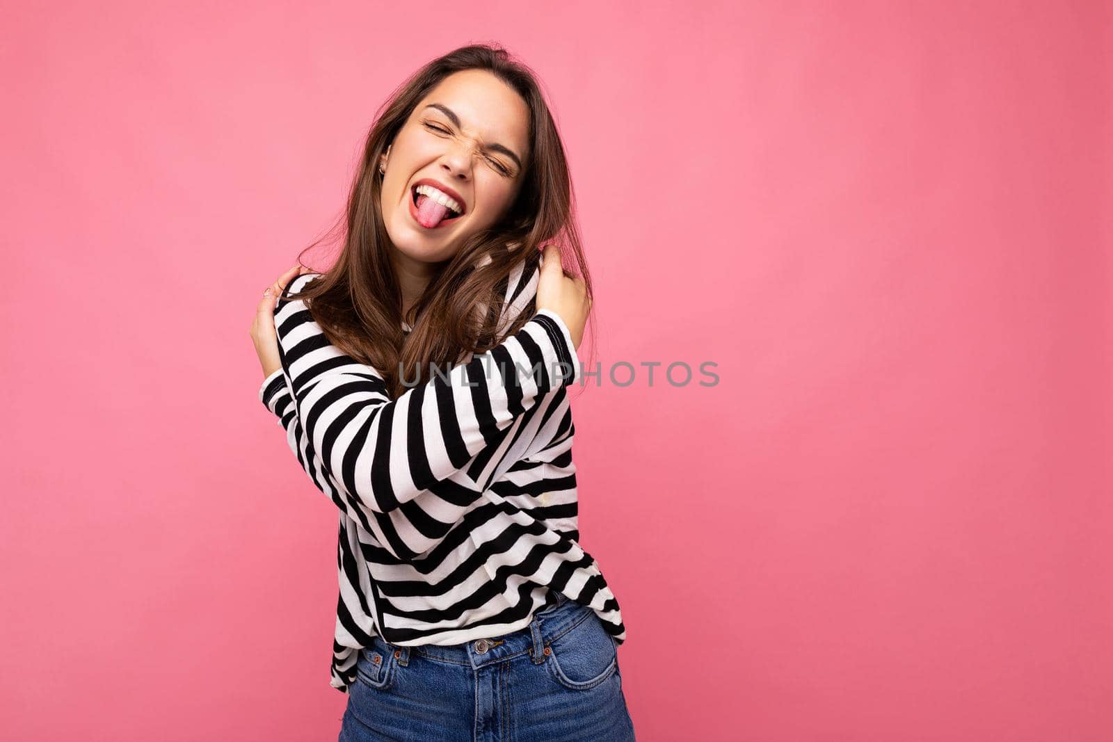 Photo shot of cute attractive pretty young brunette woman wearing casual striped longsleeve isolated over colorful background with copy space having fun and showing tongue. Positive concept