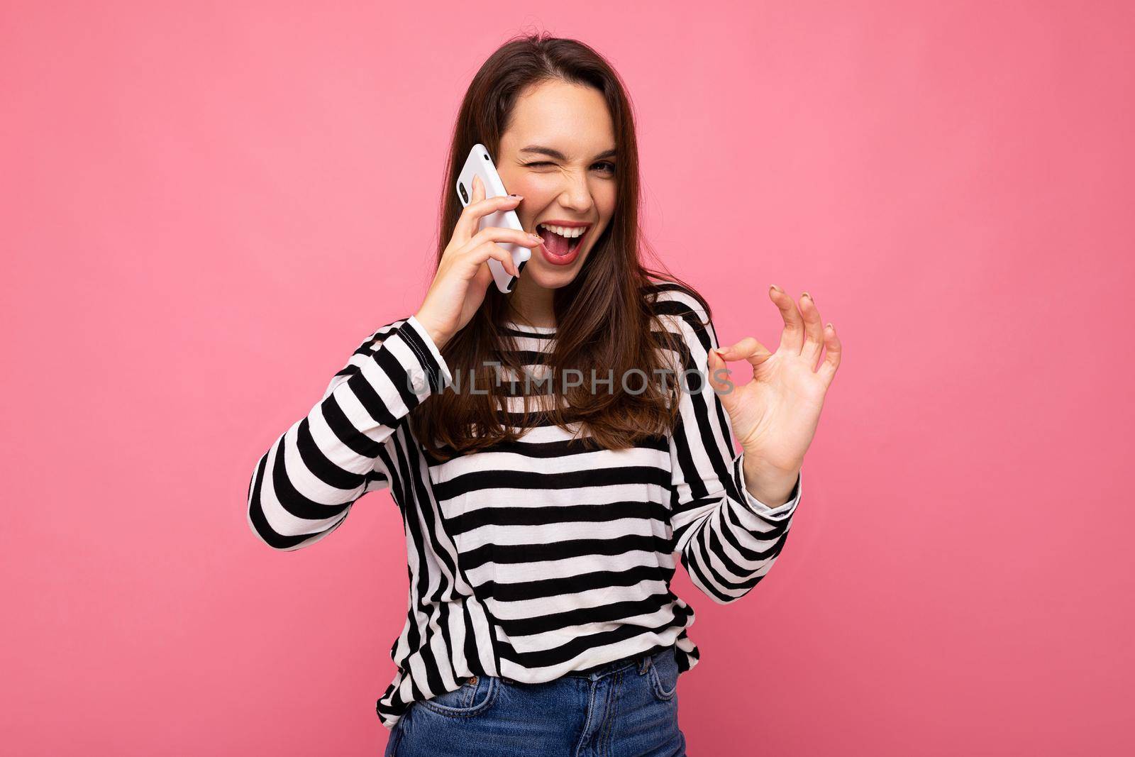 Winking Pretty happy young woman speaking on the phone wearing striped sweater isolated over background with copy space showing ok gesture looking at camera.