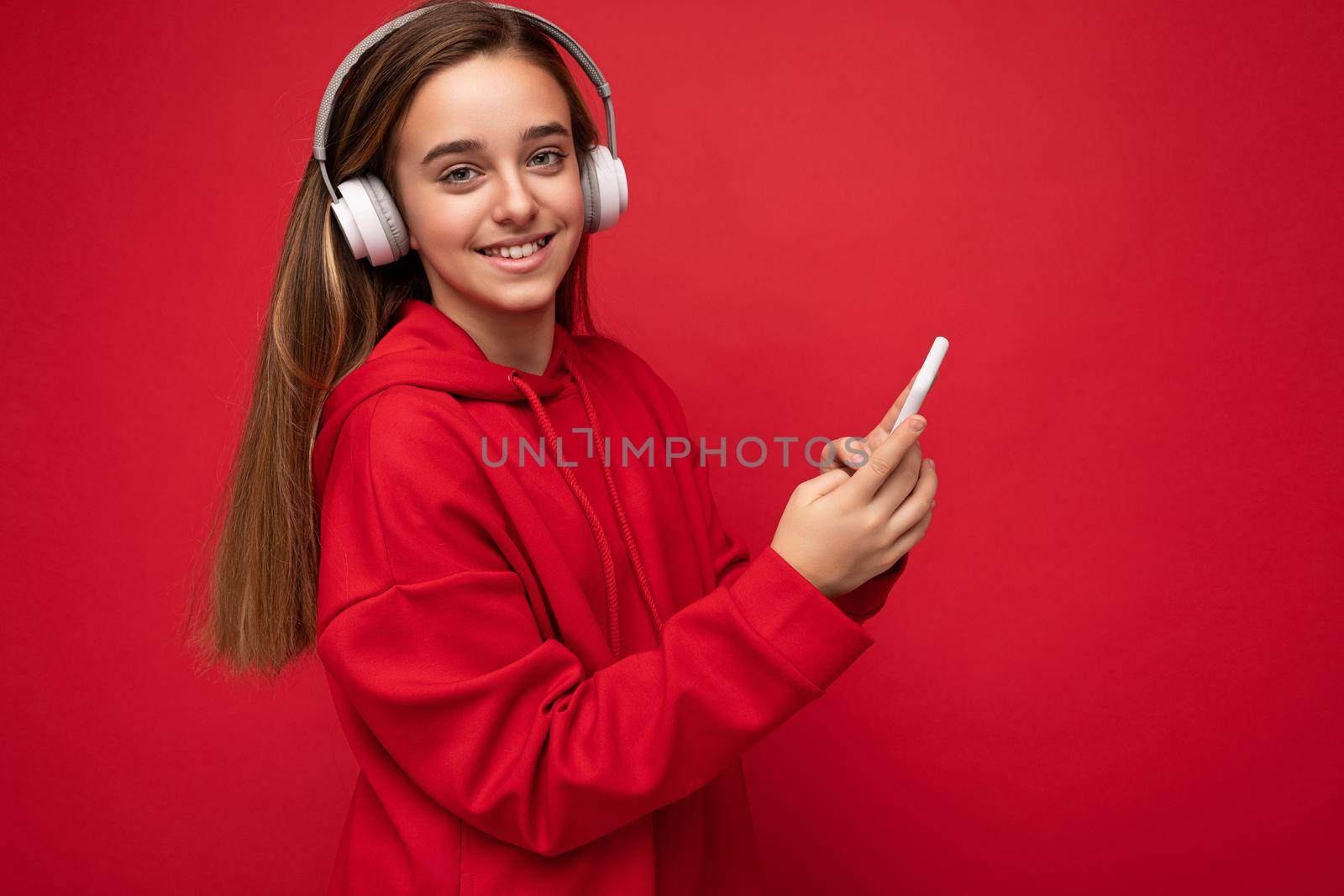 Side-profile Shot of positive smiling pretty brunette girl wearing red hoodie isolated on red background holding and using smartphone writing sms message wearing white wireless headphones listening to cool music looking at camera. empty space
