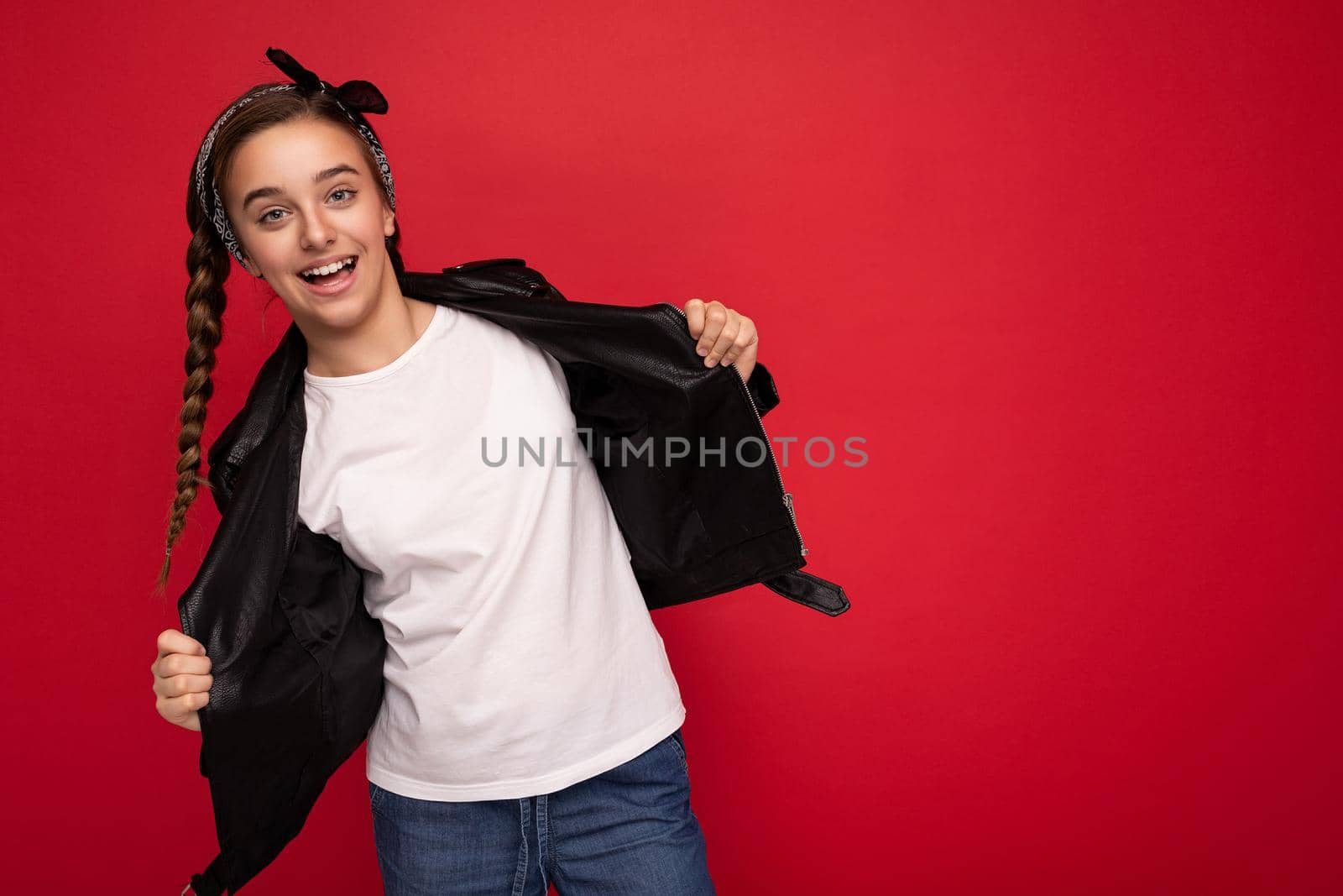 Photo shot of beautiful happy positive smiling brunette girl with pigtails wearing bandanna stylish black leather jacket and white t-shirt for mockup isolated over red background wall looking at camera. Empty space, copy space