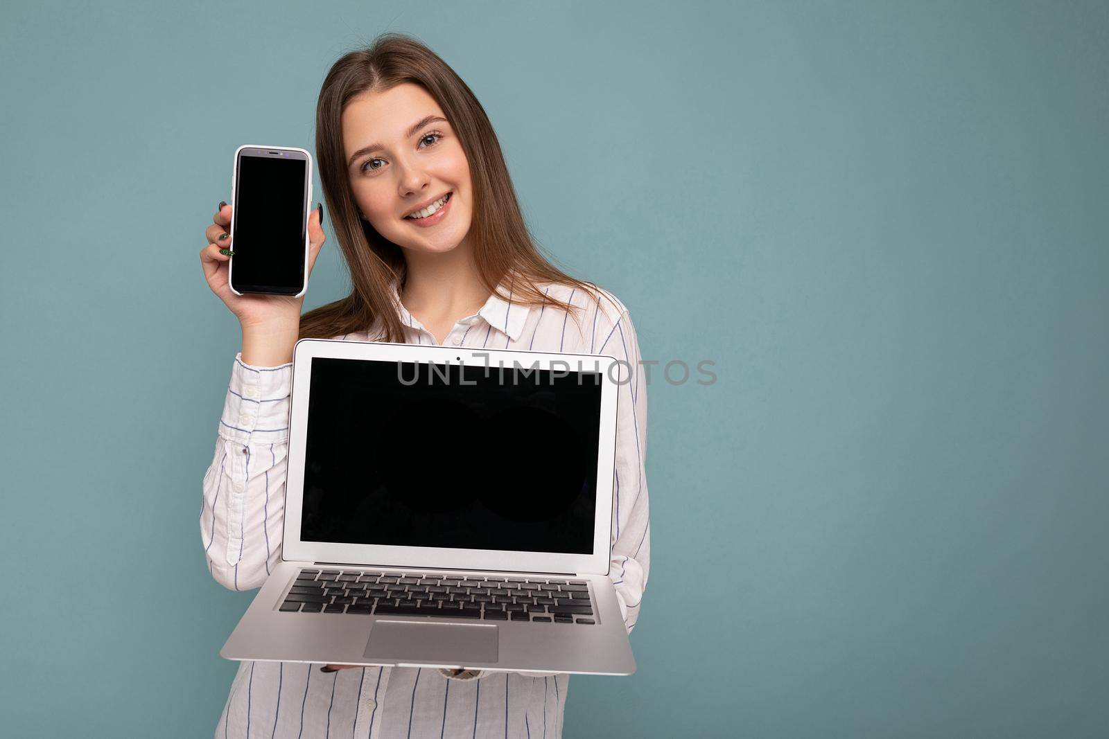 Smiling Beautiful satisfied happy young woman holding computer laptop and mobile phone looking at camera wearing shirt isolated over blue wall background. copy space, cutout