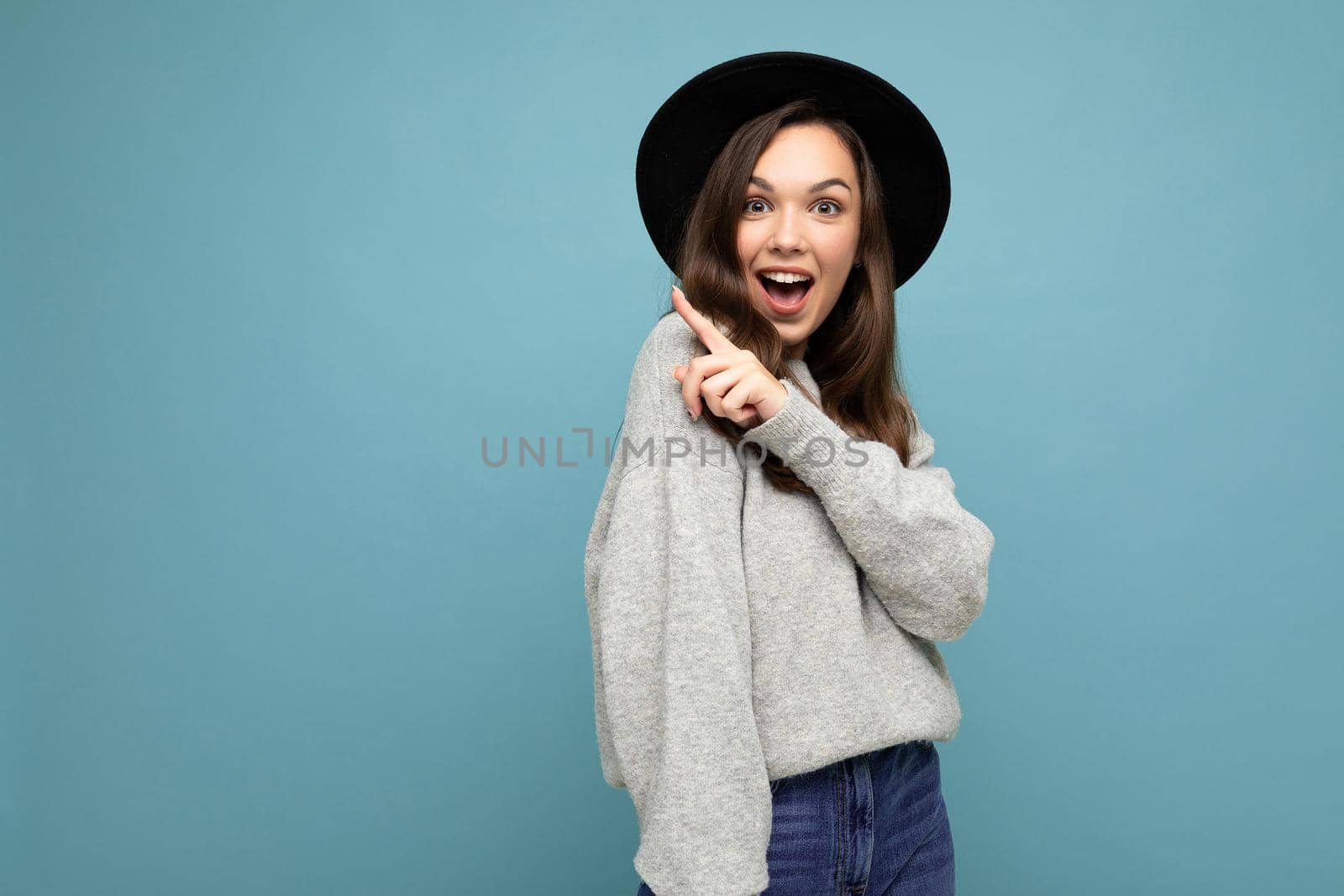 Photo of attractive brunette happy joyful young woman pointing finger at free space for text wearing grey jersey and black hat isolated on blue background.