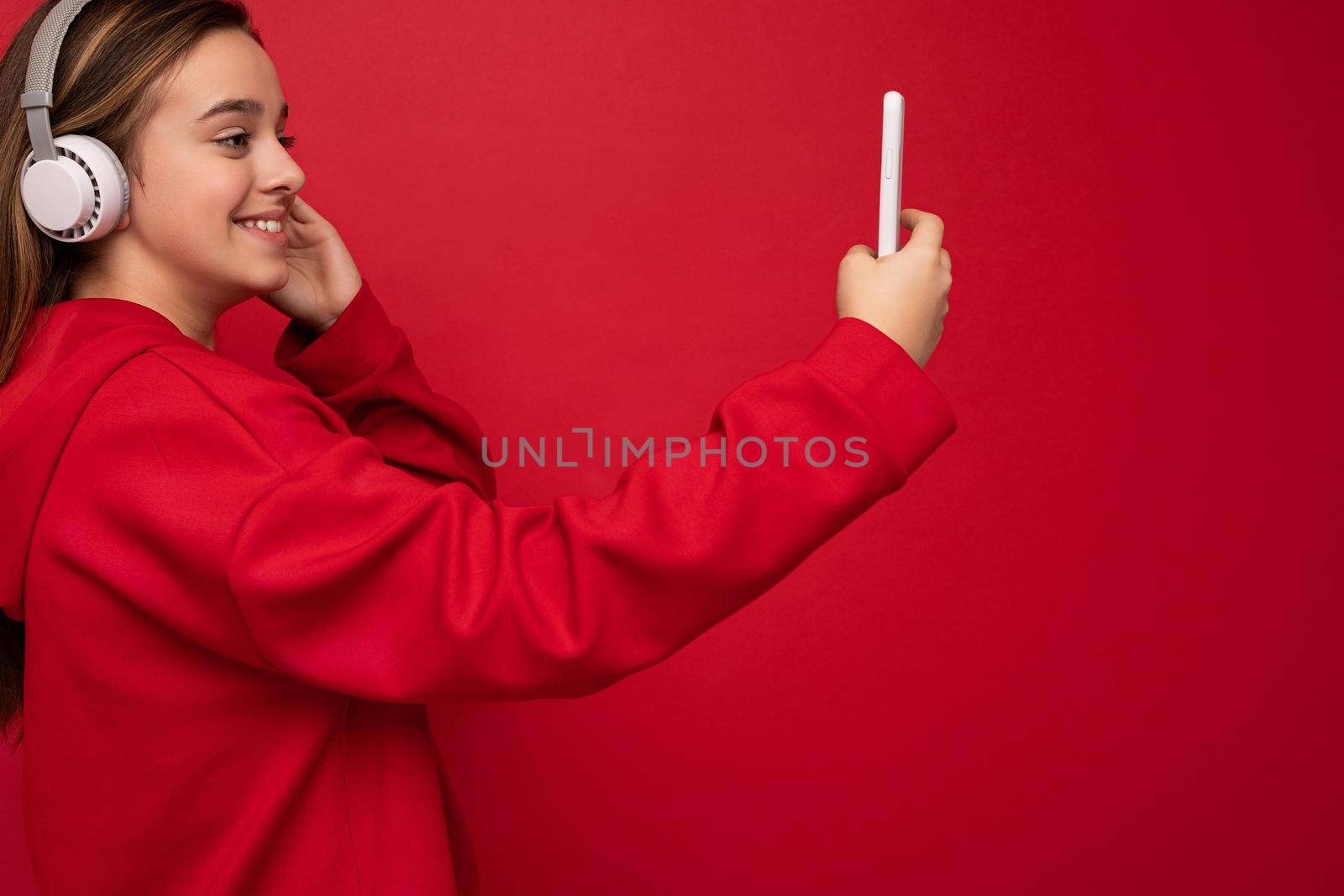 Closeup Side-profile Shot of happy smiling pretty brunette girl wearing red hoodie isolated on red background holding and using smartphone taking selfie photo wearing white wireless headphones listening to cool music looking at phone display. empty space