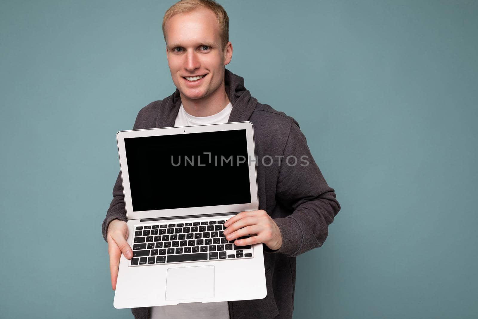 Close-up portrait of handsome smiling blonde man holding computer laptopwith empty monitor screen with mock up and copy space looking at camera isolated over blue background.