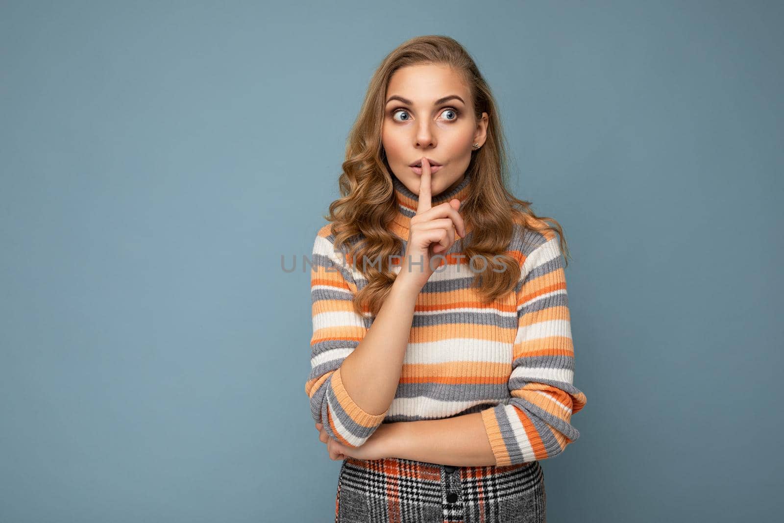 Shot of young blonde wavy-haired woman with sincere emotions wearing striped sweater isolated on blue background with copy space and showing shhh gesture. Keep secret.