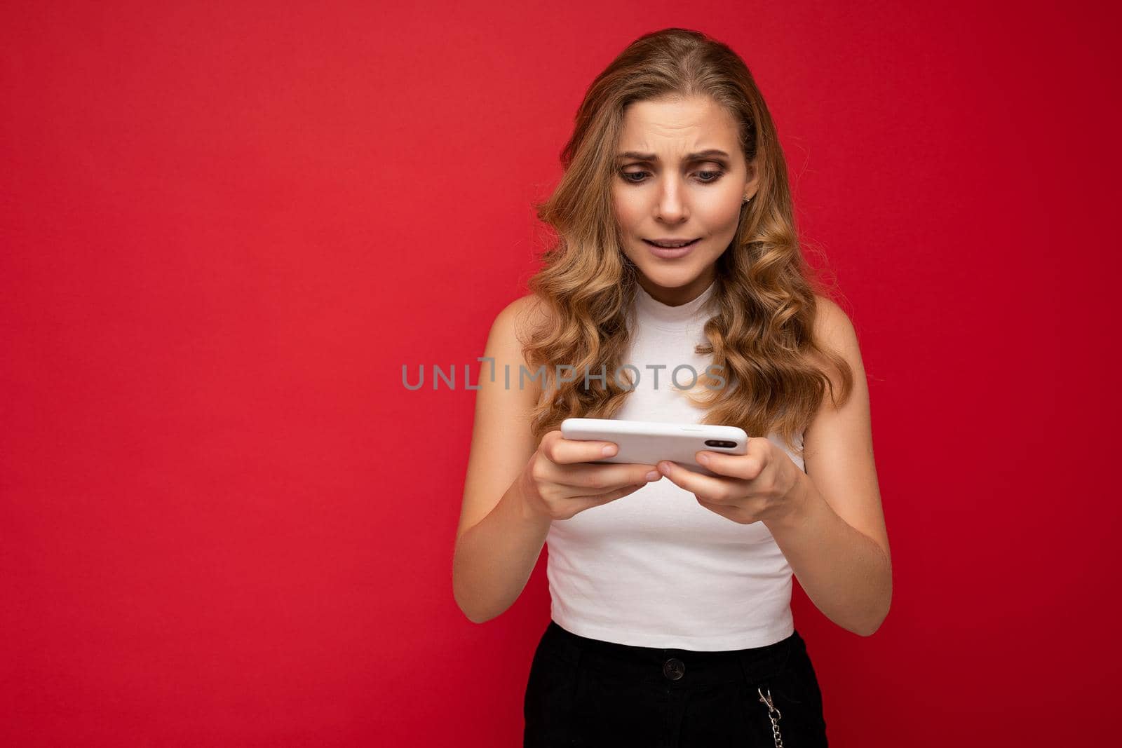 Photo of concentrated beautiful young blonde woman wearing white t-shirt isolated on red background using smartphone and playing phone games looking at gadjet screen.