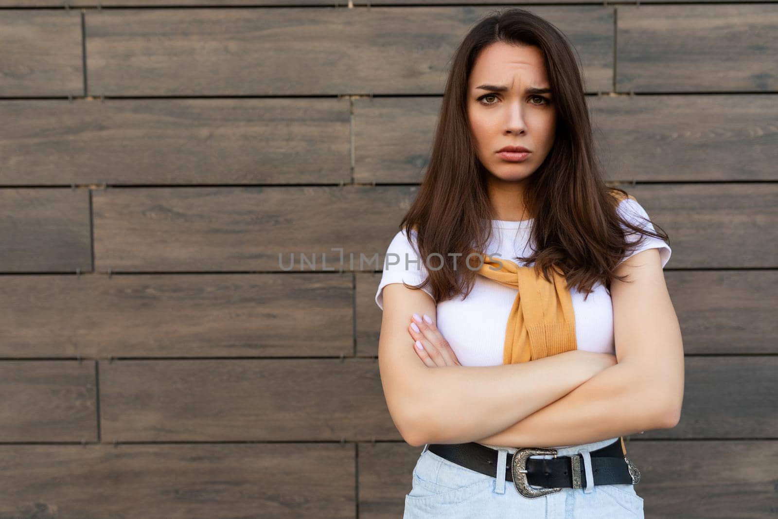 Photo of touchy sad upset offended resentful young brunet woman wearing casual white t-shirt and jeans with yellow sweater poising near brown wall in the street.