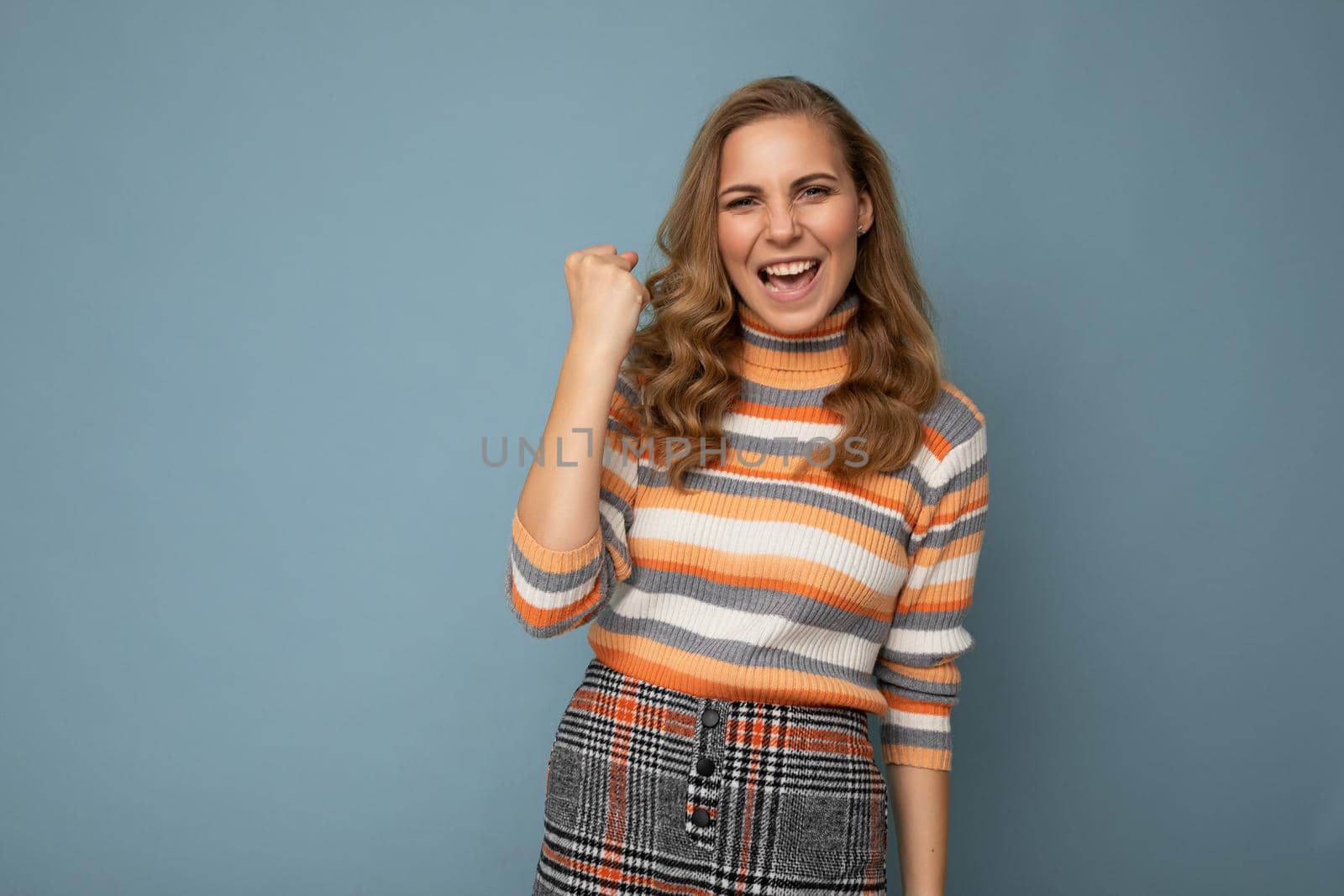 Portrait of happy positive young attractive blonde curly woman with sincere emotions wearing trendy striped pullover isolated on blue background with copy space and celebrating victory saying yeah.