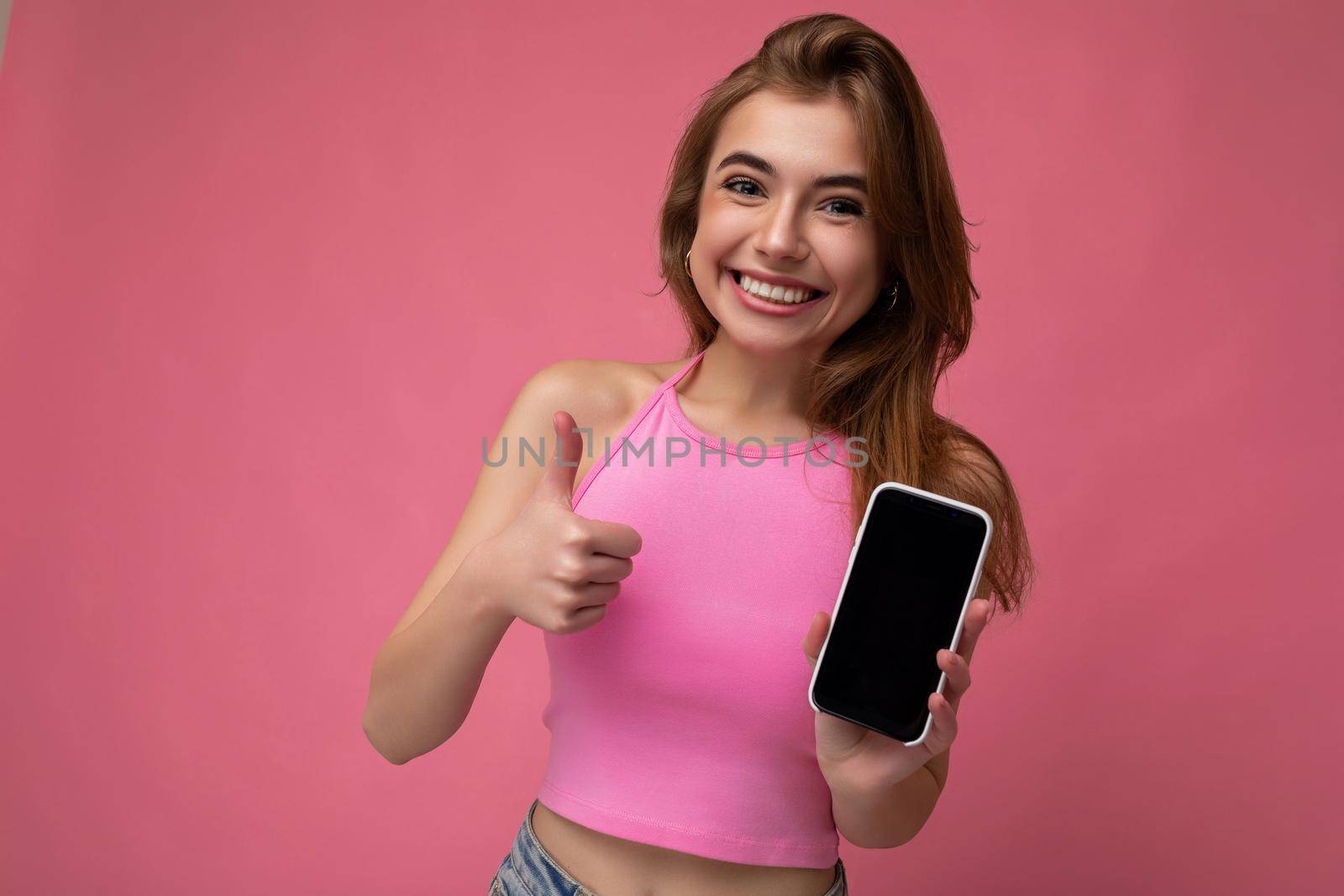 Beautiful joyful young blonde woman wearing pink top poising isolated on pink background with empty space holding in hand and showing mobile phone with empty display for mockup looking at camera and showing thumbs up gesture.