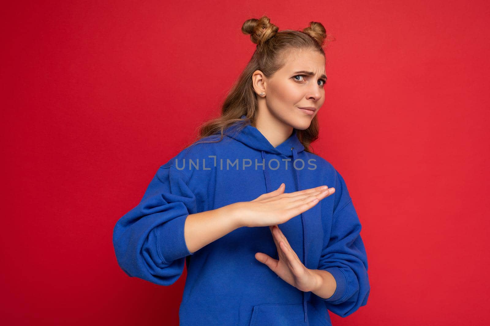 Photo of emotional dissitisfied young pretty blonde woman with two horns with sincere emotions wearing hipster bright blue hoodie isolated over red background with copy space and showing timeout gesture.