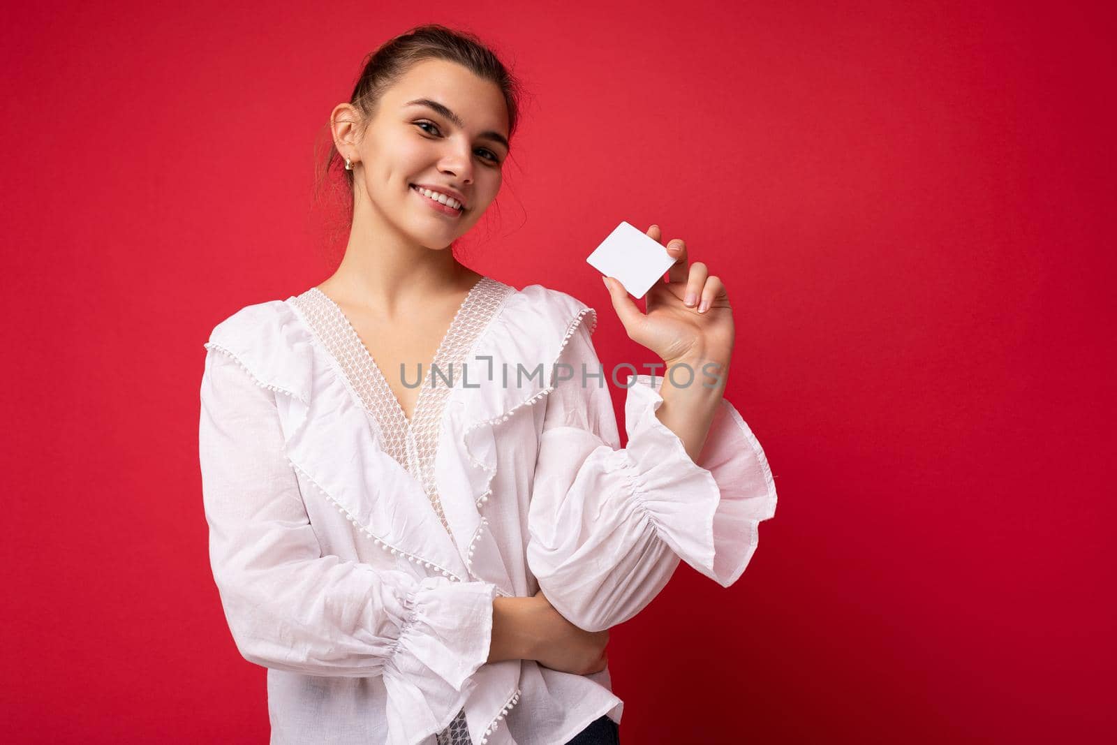 Photo of beautiful positive young dark blonde woman wearing white blouse isolated over red background holding credit card looking at camera.