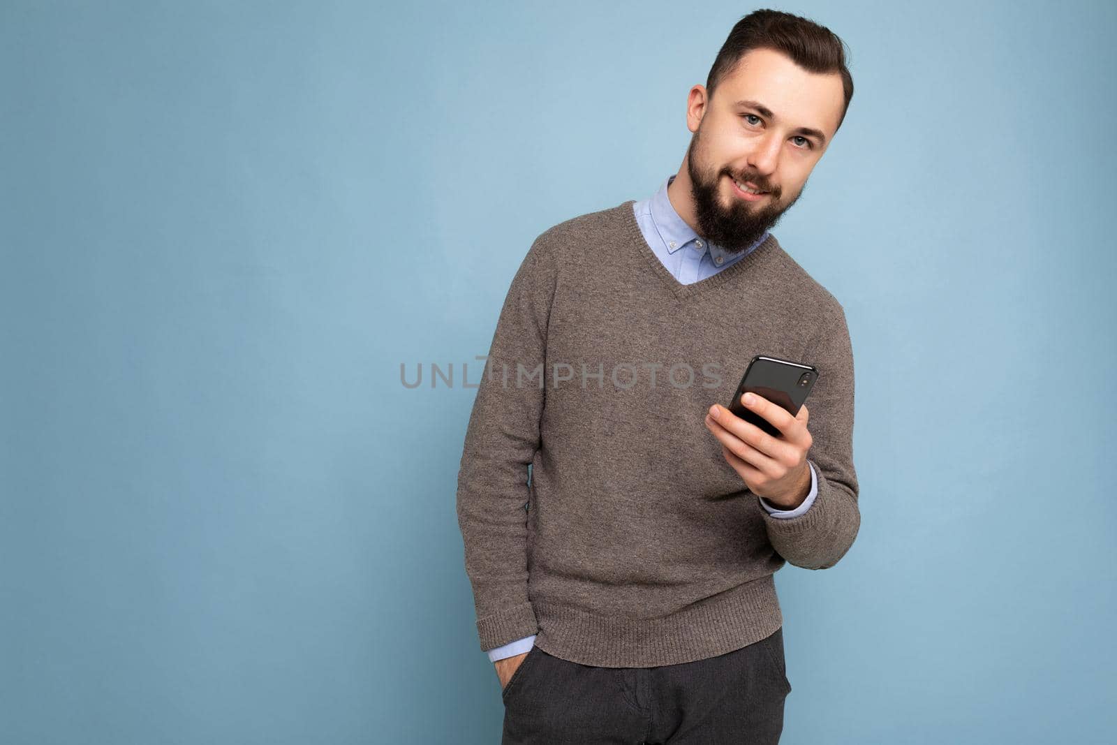 Photo of handsome good looking brunet bearded young man wearing grey sweater and blue shirt isolated on pink background with empty space holding in hand and using mobile phone communicating online looking at camera.