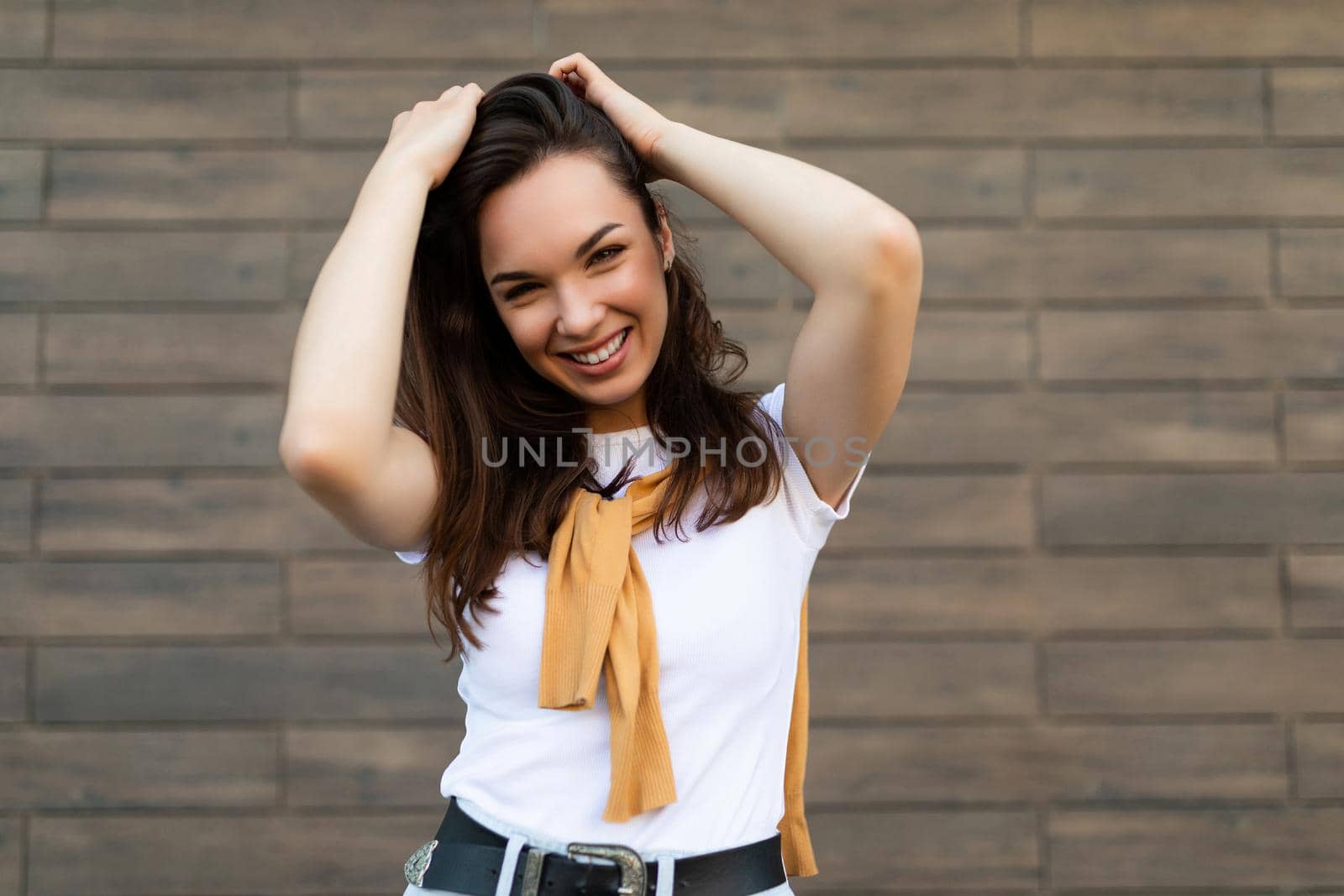 Portrait of successful smiling joyful happy young brunet woman wearing casual white t-shirt and jeans with yellow sweater poising near brown wall in the street and having fun.